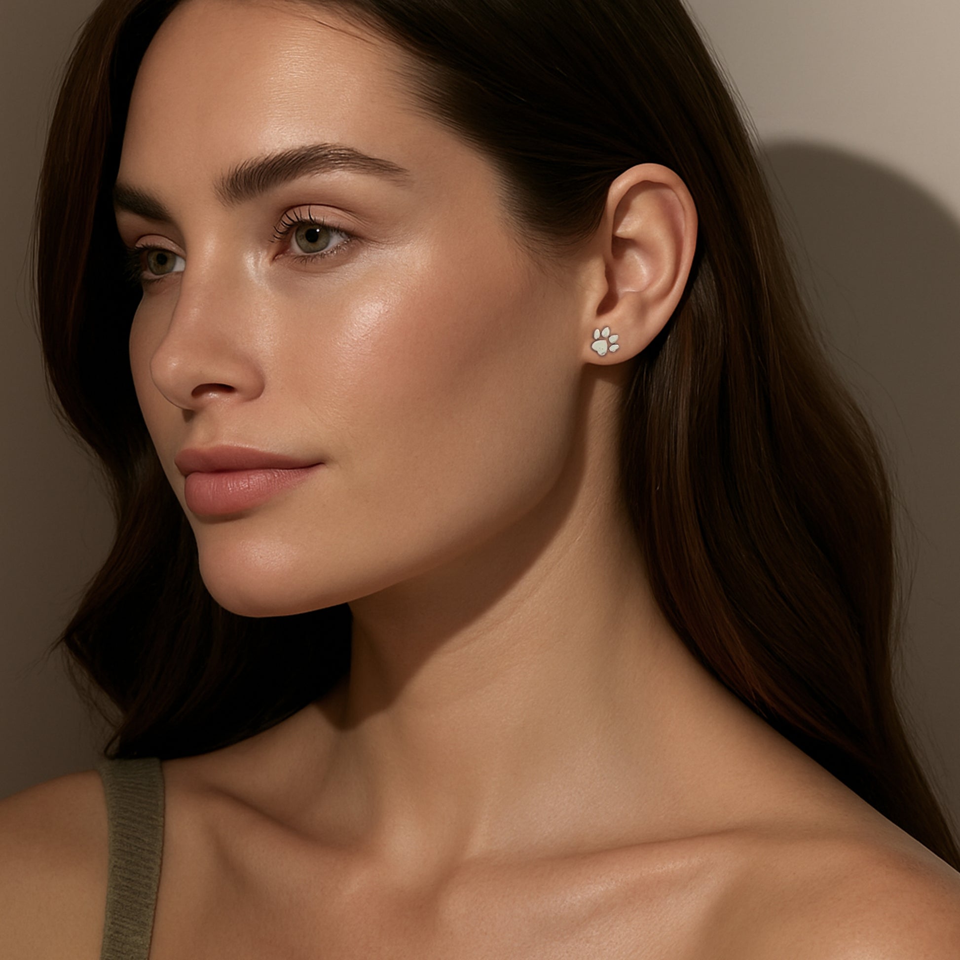  A woman with long brown hair and soft makeup is shown in side profile wearing a small silver paw-shaped stud earring. She is dressed in a green tank top, and the lighting creates gentle shadows against a neutral background, emphasizing her natural beauty and the earring.