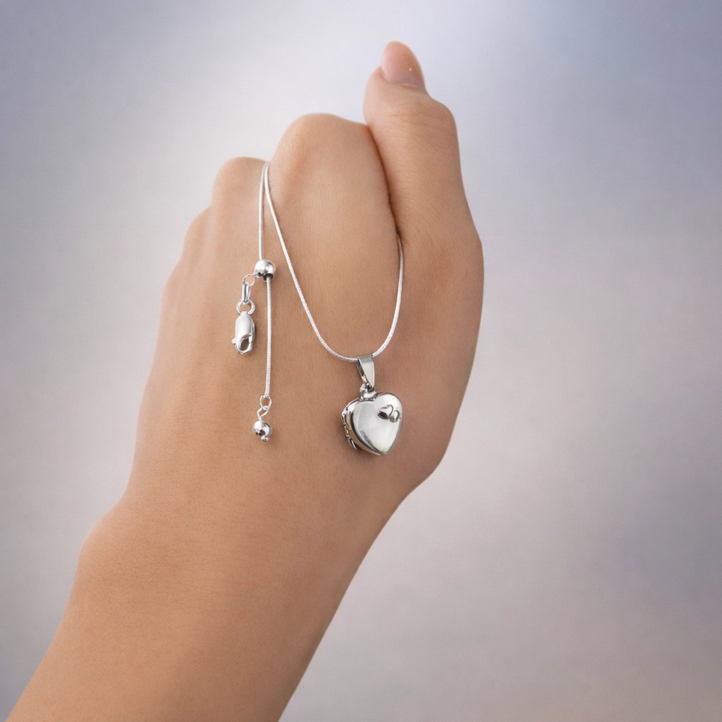 Polished silver heart locket necklace on a delicate snake chain, displayed on a hand, with adjustable bolo bead and smooth studio background.