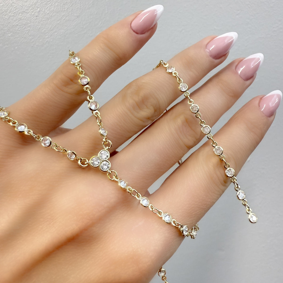 Close-up of a hand with pink and white French manicure holding a sparkling gold lariat necklace with bezel-set round cubic zirconia stones, showcasing its delicate and elegant design.