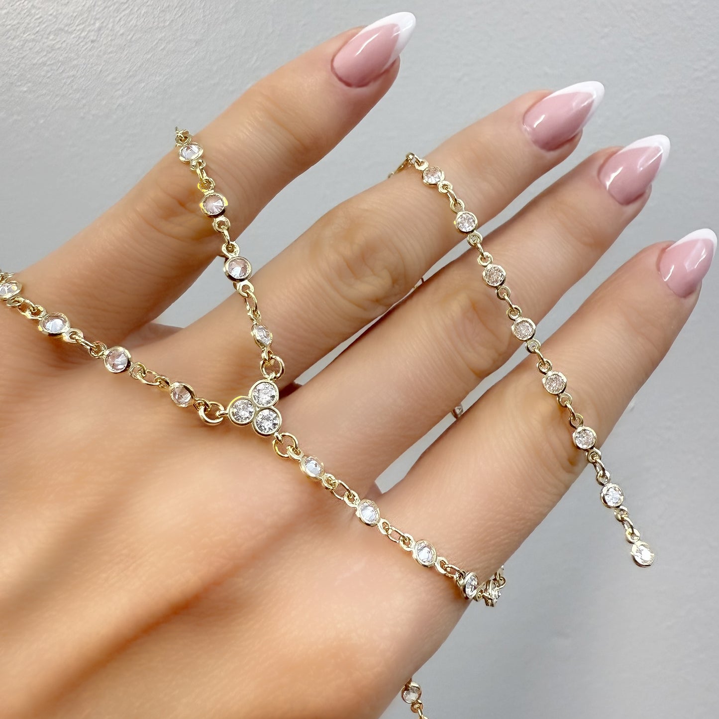 Close-up of a hand with pink and white French manicure holding a sparkling gold lariat necklace with bezel-set round cubic zirconia stones, showcasing its delicate and elegant design.