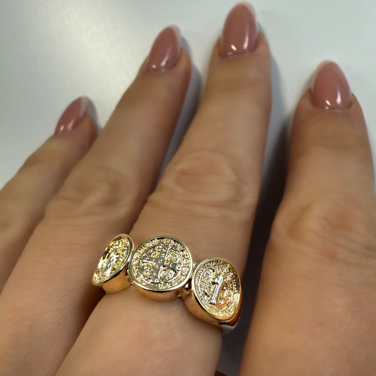 Close-up of a woman's hand wearing a bold gold-filled St. Benedict ring with three oval medallions featuring intricate saint and cross engravings, set on a polished band against a neutral background.