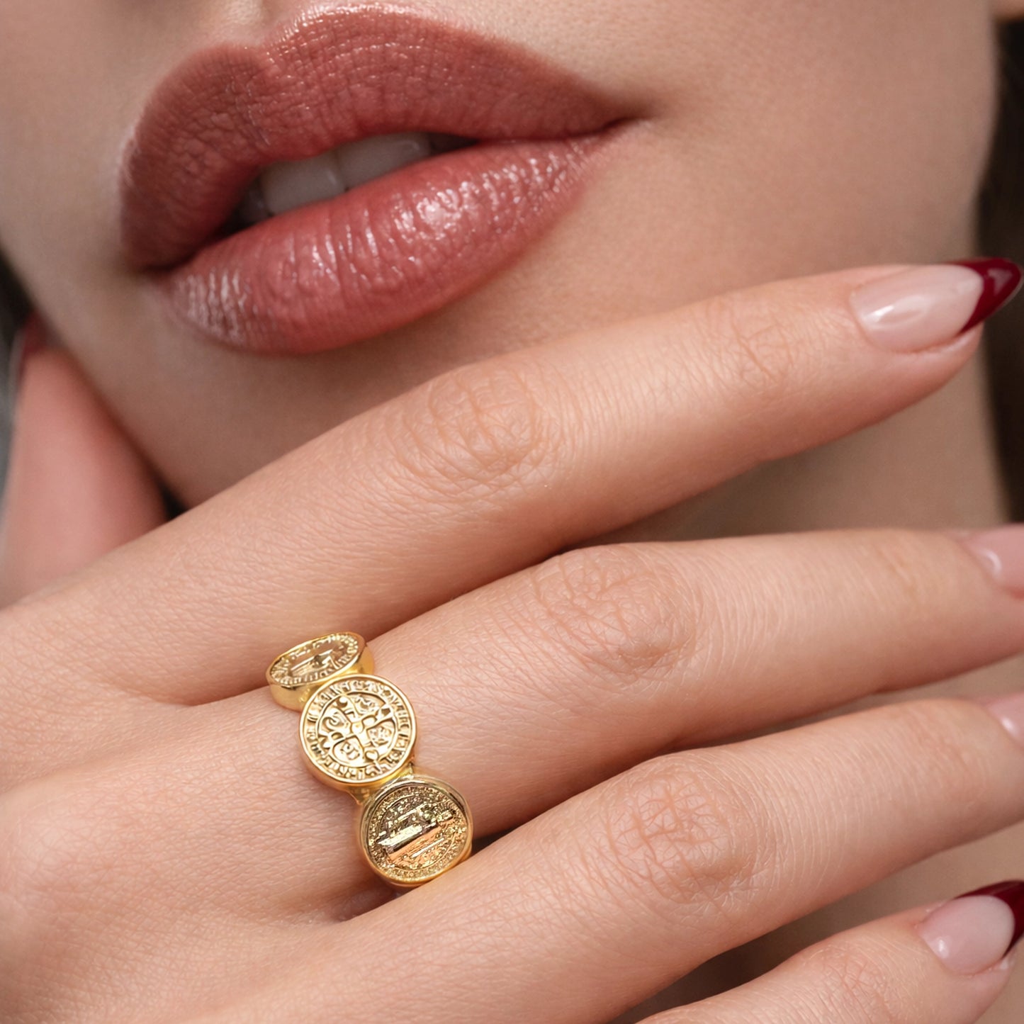 Gold Saint Benedict ring with three round medallions on a woman’s hand, close-up studio shot showing detailed engravings, polished finish, natural skin tone, elegant manicure, and soft neutral background.
