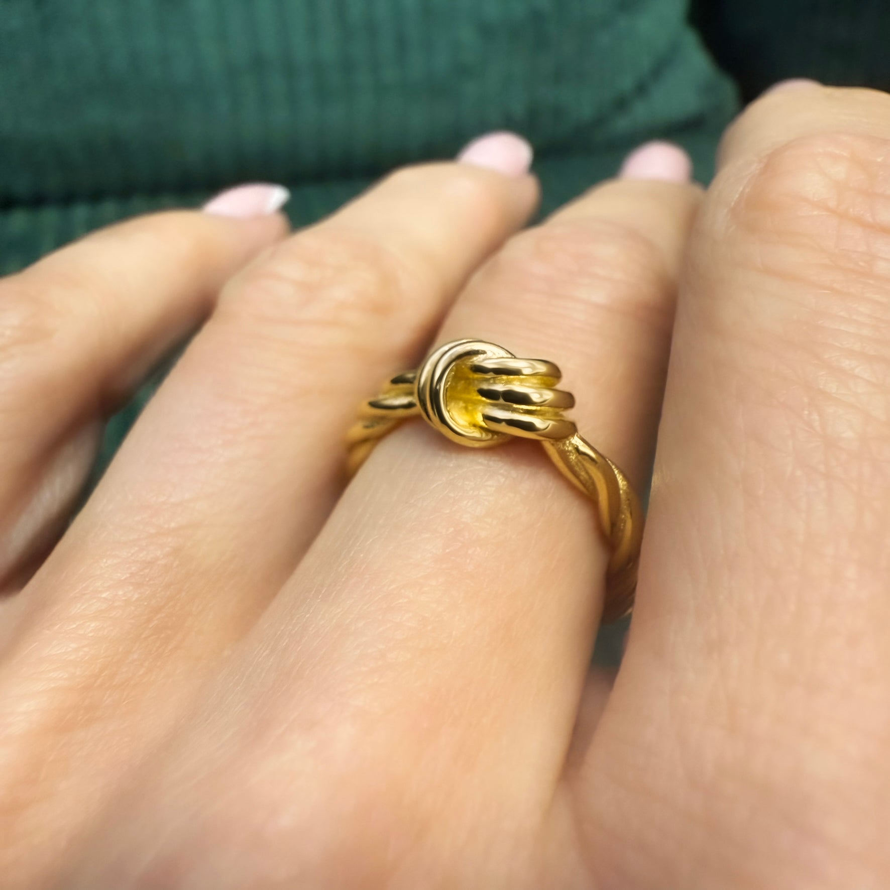 Close-up of a woman's hand wearing a gold plated knot ring with a thick twisted band and layered rope-style center. The ring has a shiny finish and is worn over a natural pink French manicure, with a dark green background.