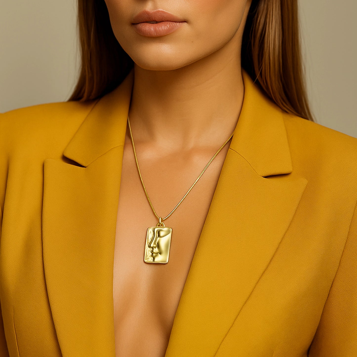 Model wearing a bold matte gold plated abstract face pendant necklace on a sleek snake chain, styled with a mustard yellow blazer. The statement rectangular charm features modern, artistic facial lines, creating an elegant and contemporary look.