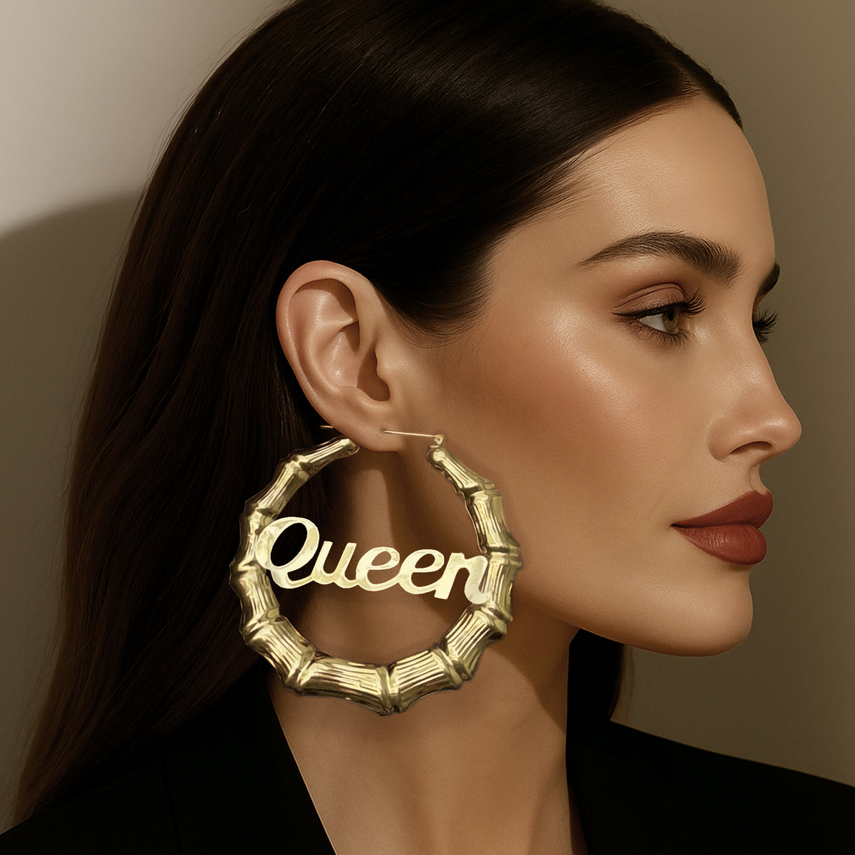 Side profile of a woman wearing oversized gold-tone bamboo hoop earrings with the word "Queen" in bold script, styled with sleek hair and bold makeup.