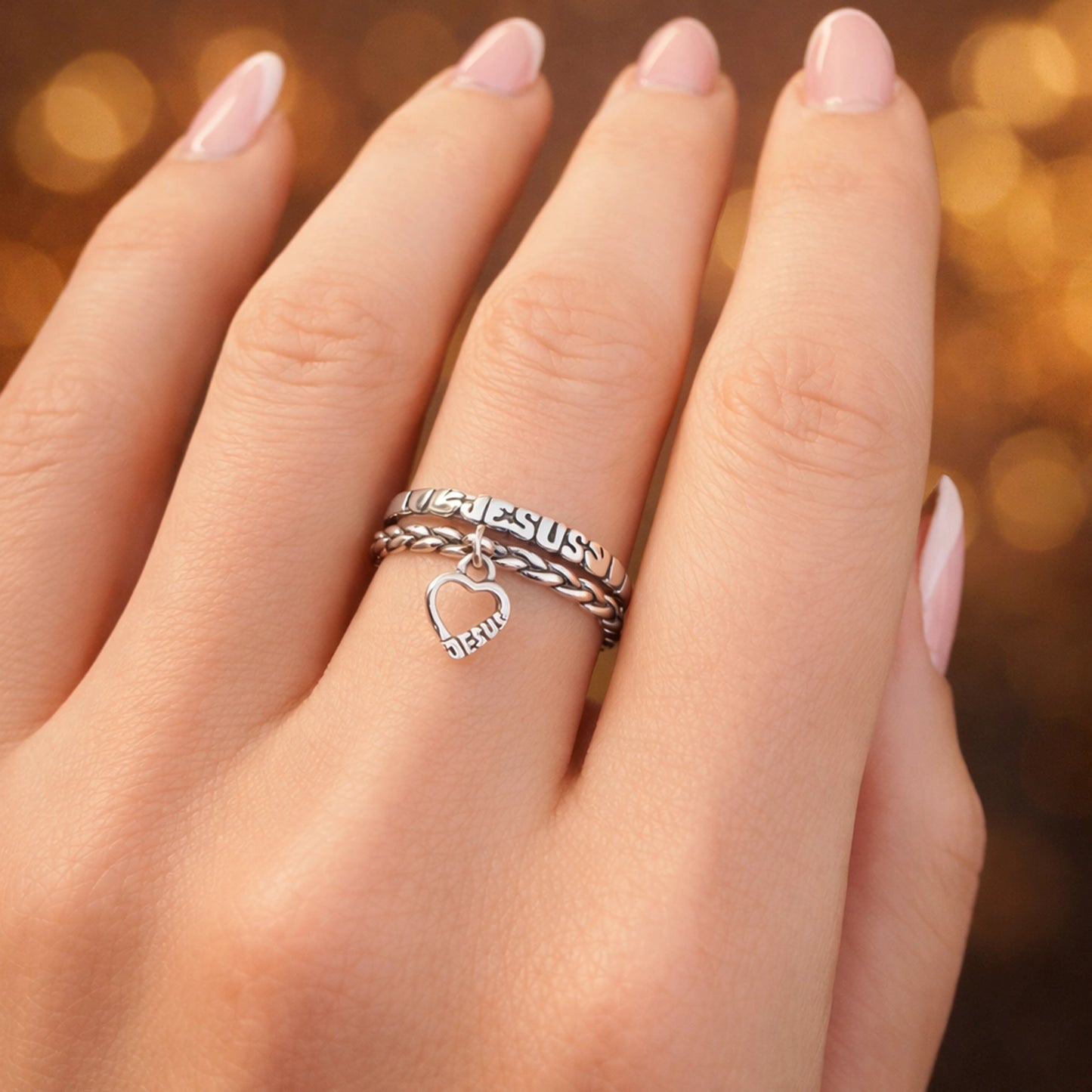 Sterling silver Jesus stackable ring set with braided band and dangling heart charm engraved “Jesus,” worn on hand with French manicure, Christian faith jewelry ring.