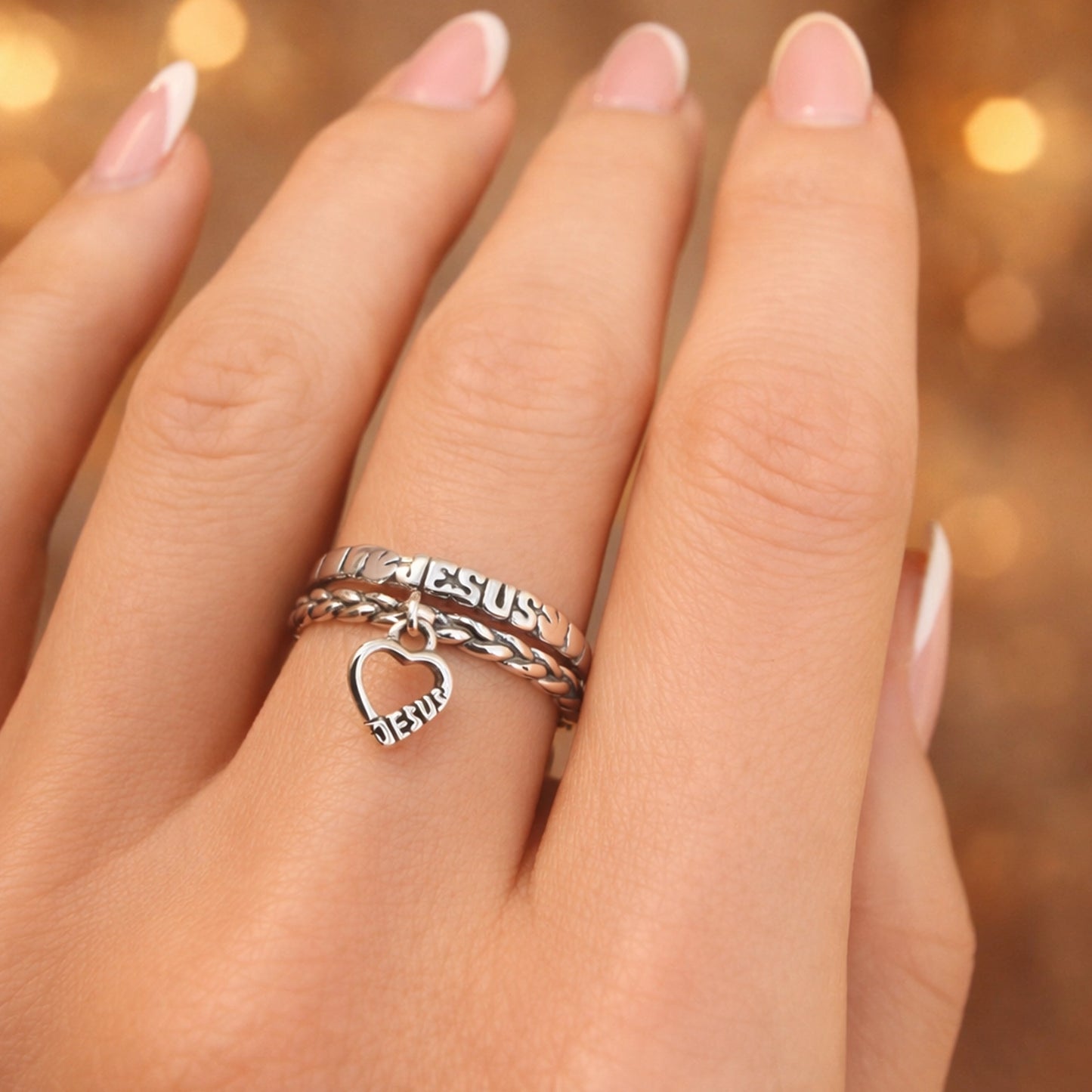 Sterling silver Jesus stackable ring set with braided band and dangling heart charm engraved “Jesus,” worn on hand with French manicure, Christian faith jewelry ring.