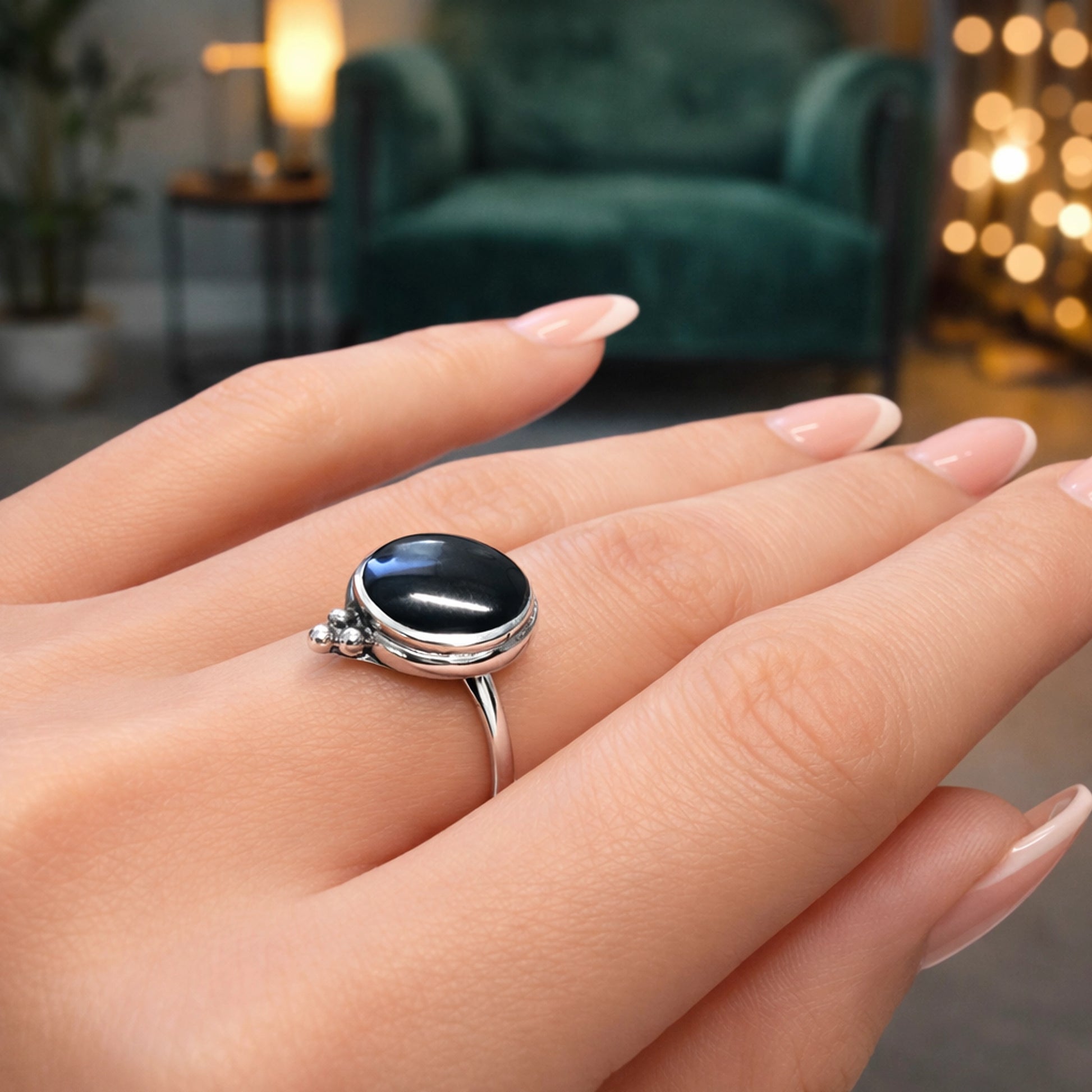 Close-up of a woman’s hand wearing a minimalist silver ring with an oval black cabochon stone set in a smooth bezel setting. The ring features a slim polished band and small decorative silver bead accents on one side of the setting. The model has nude almond-shaped manicured nails, and the background shows a softly blurred green armchair and warm ambient lights, creating a cozy indoor atmosphere.