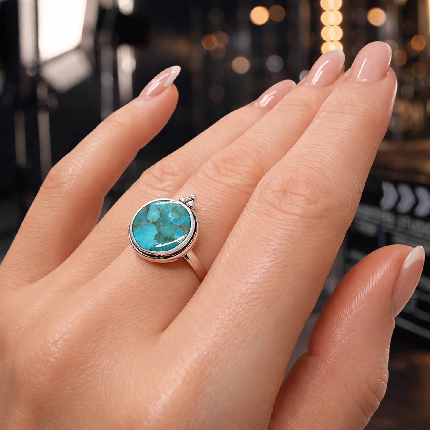 Close-up of a woman’s hand wearing a round turquoise statement ring set in a polished silver bezel on the ring finger. The turquoise stone features natural blue-green marbling with subtle veining. Her nails are almond-shaped with glossy nude-pink polish. The background is softly blurred with warm studio lighting and bokeh highlights, creating a professional jewelry photography look.