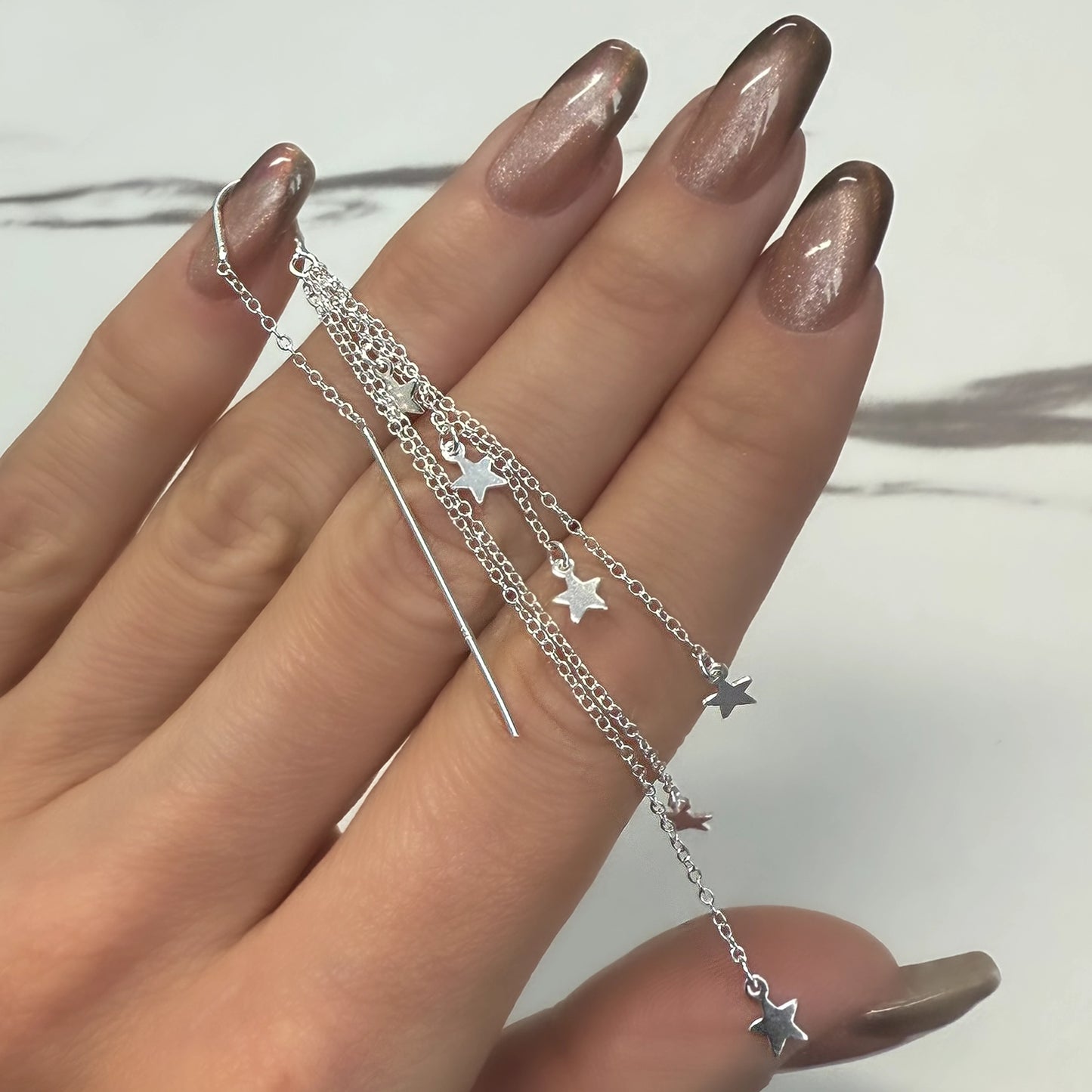 A close-up of a hand with almond-shaped metallic rose-gold and black gradient nails holding a pair of delicate sterling silver threader earrings. The earrings feature fine chains adorned with small, dangling star charms, creating a celestial and whimsical look. The white marble-style background adds contrast to the polished silver and sparkling nail finish.