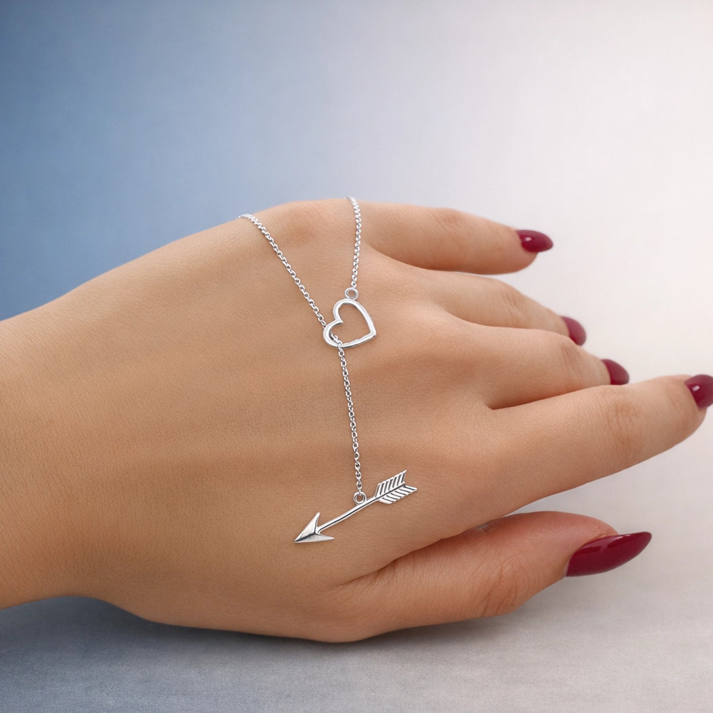 Sterling silver hand chain bracelet with open heart charm and arrow detail, shown on a woman’s hand, polished finish with delicate chain, minimal studio background.