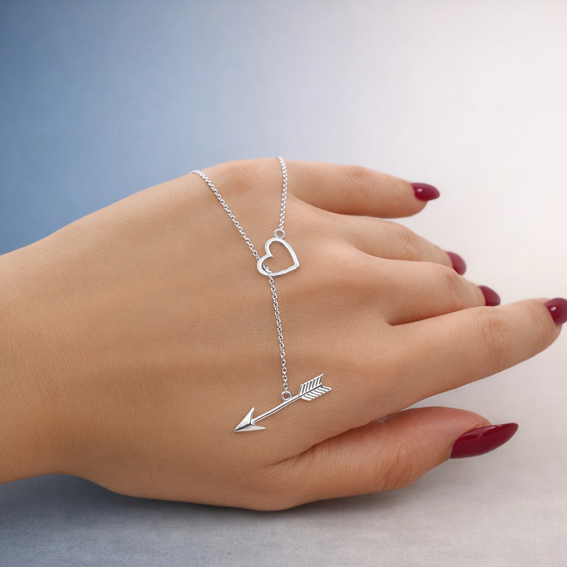 Sterling silver hand chain bracelet with open heart charm and arrow detail, shown on a woman’s hand, polished finish with delicate chain, minimal studio background.
