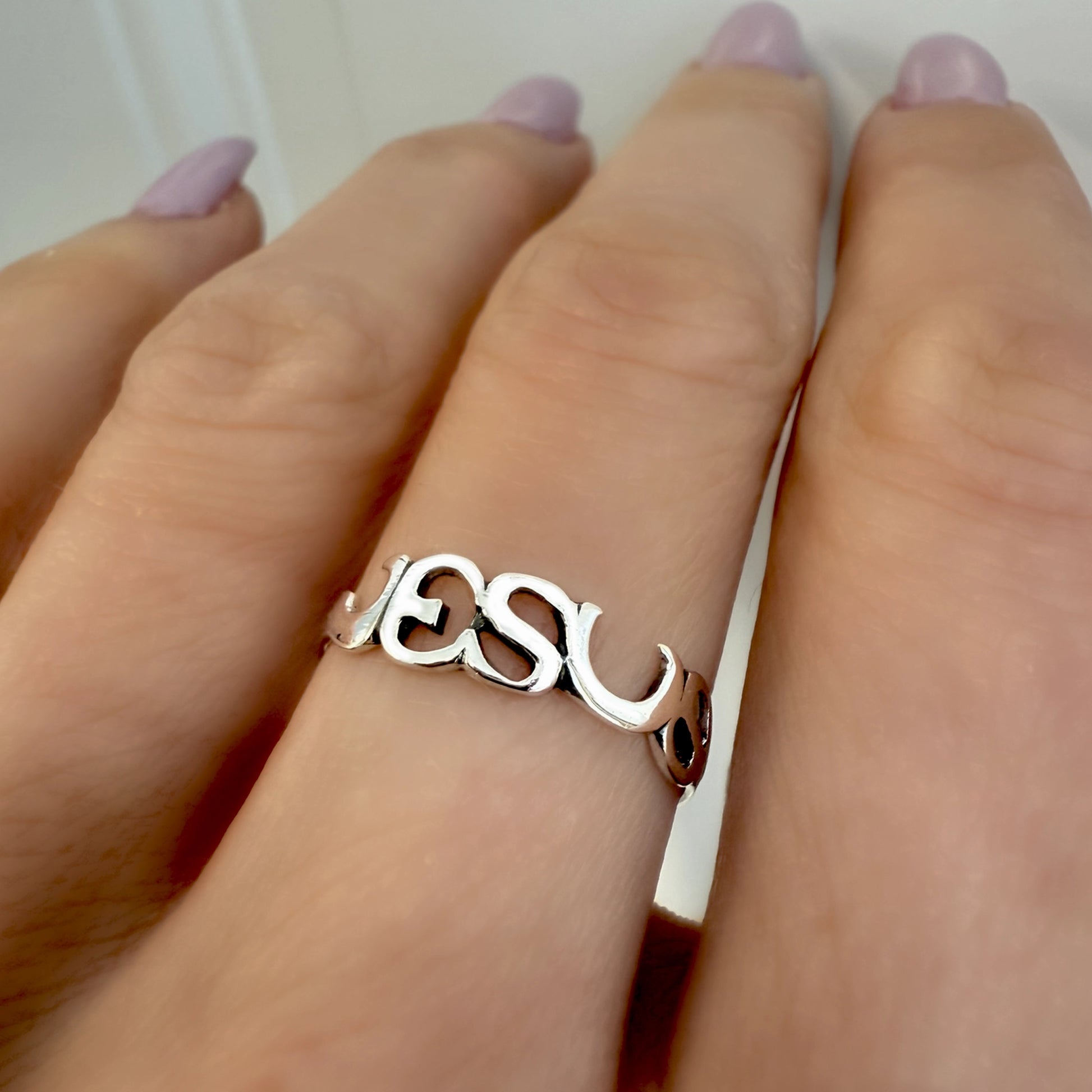 Sterling silver cut-out ring spelling “JESUS” worn on a finger with light purple nails.