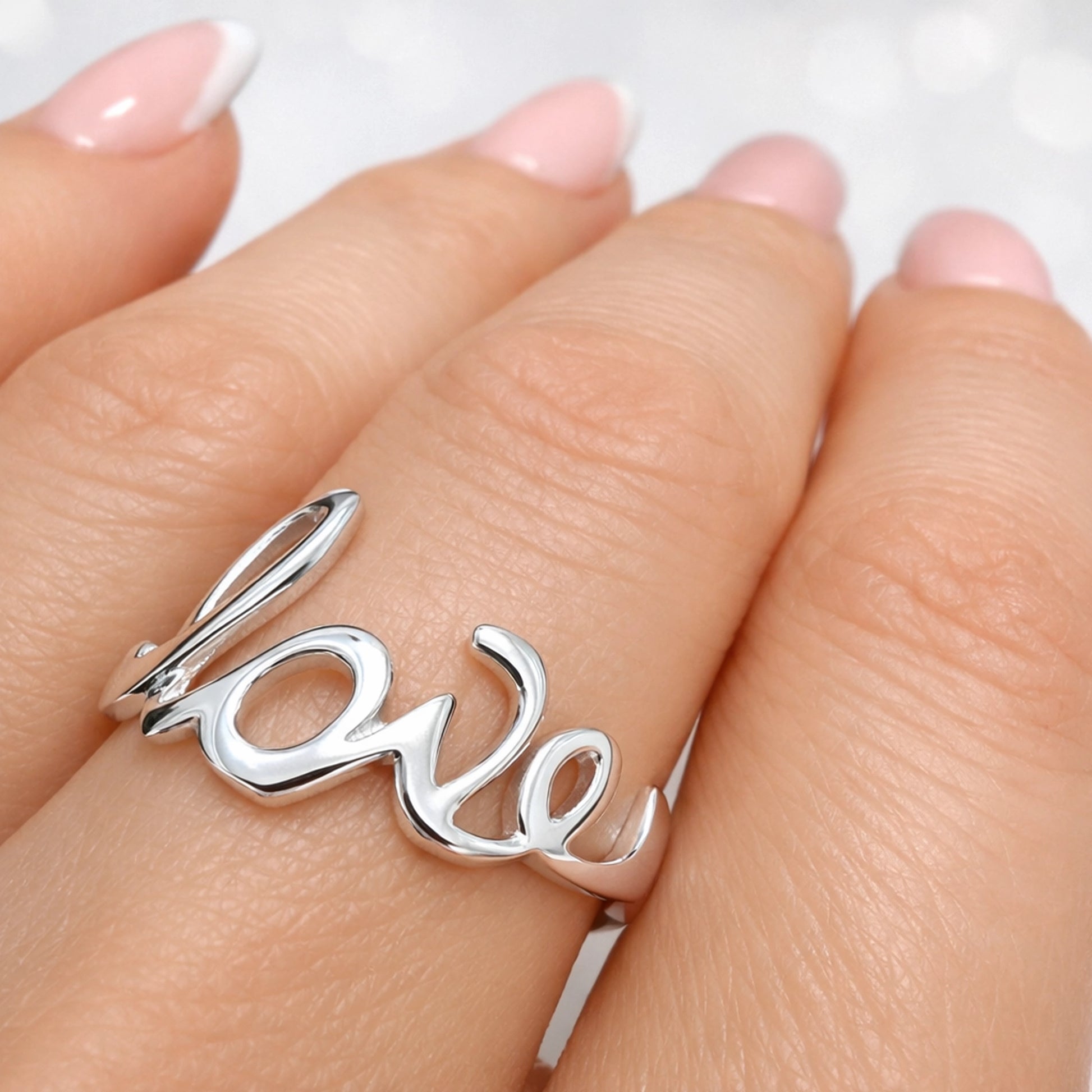 Sterling silver cut-out ring spelling the word "love" in a cursive script, worn on a hand with light pink polished nails.