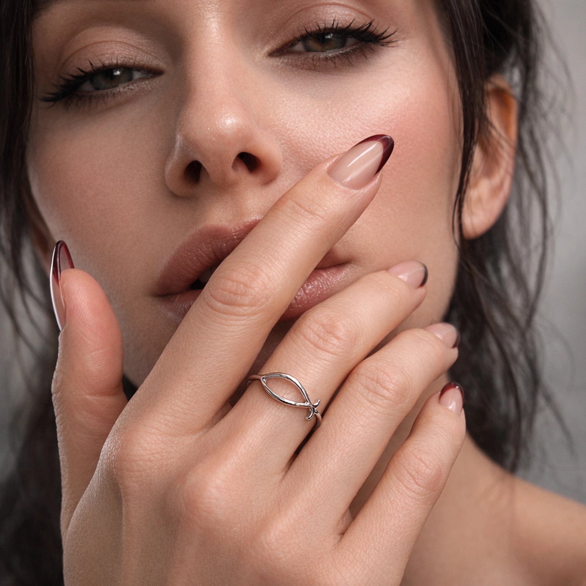 Close-up square studio portrait of a beautiful woman with natural glowing skin and soft nude lips, showing a minimalist polished silver fish symbol ring on her finger, elegant hands with a clean nude manicure, luxury jewelry presentation.