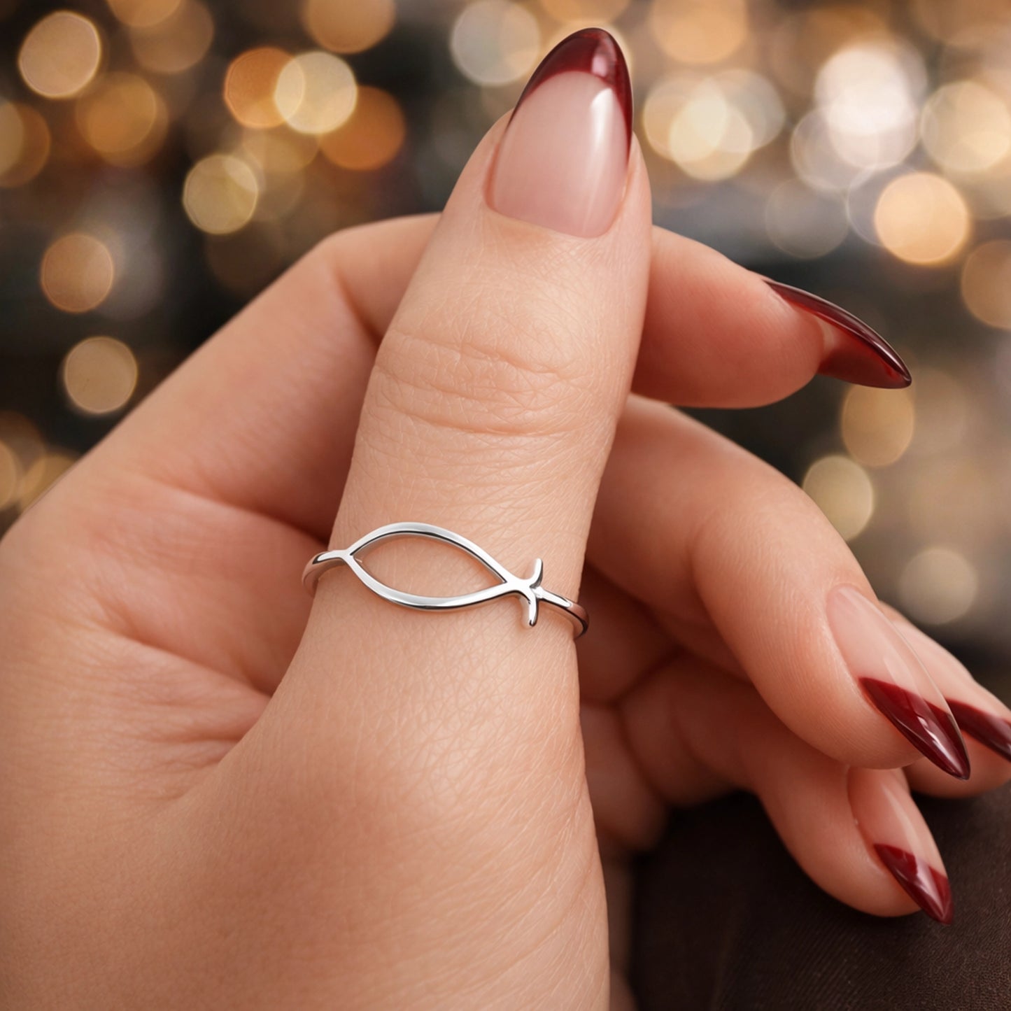 close-up of a woman’s hand wearing a polished silver Ichthys fish ring, professionally retouched with smooth natural skin, elegant red gradient manicure, dark brown trousers, and a soft studio background with warm bokeh lights, highlighting the ring’s clean design without glare.