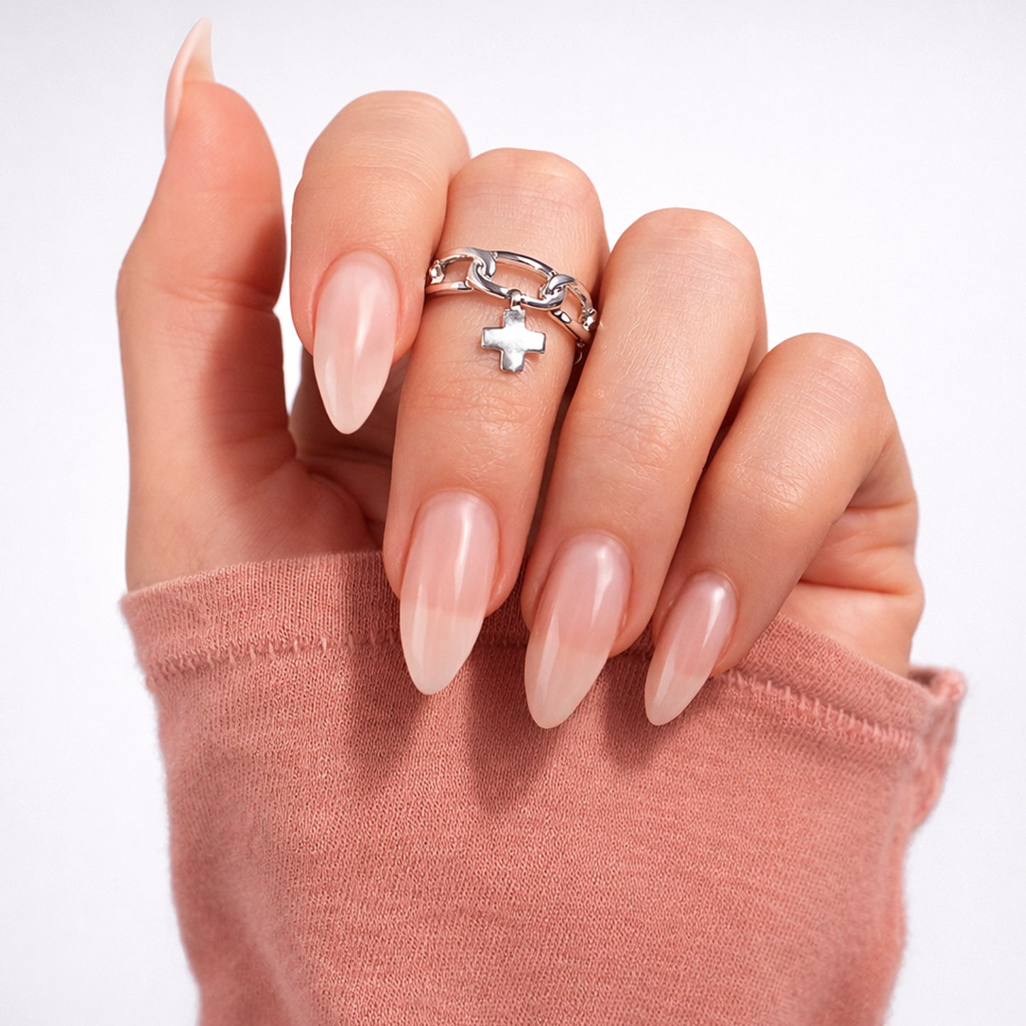 Close-up of a woman’s hand wearing a polished sterling silver chain ring with a small cross charm, shown on manicured nude almond-shaped nails against a clean white studio background.