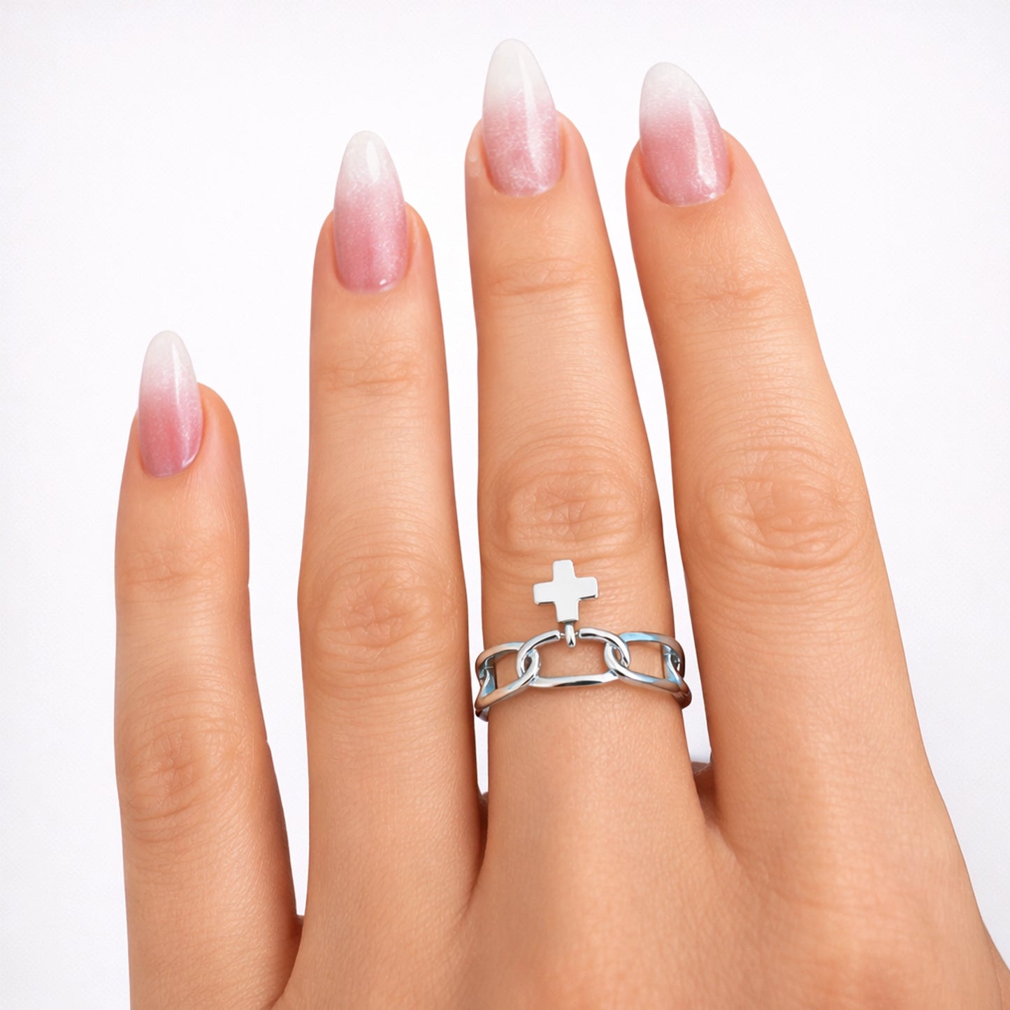 Close-up of a woman’s hand with almond-shaped pink-to-white ombre manicure wearing a polished sterling silver chain ring with a small cross charm on a clean white studio background.