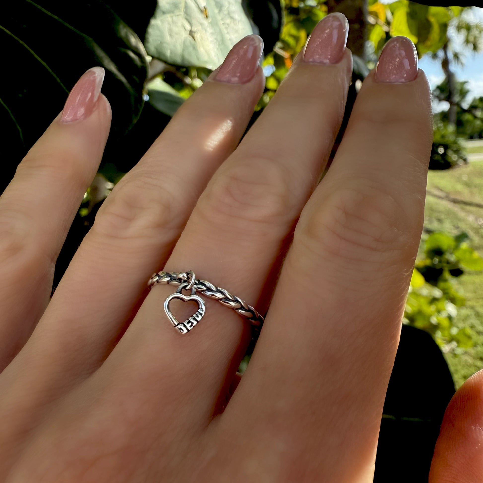 Christian charm ring with a dangling heart engraved “Jesus,” shown on a model's hand with soft pink nails.