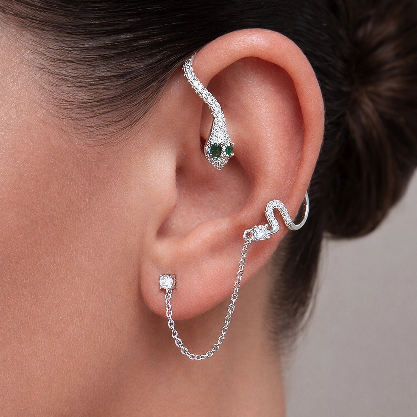 Silver snake ear jacket earring with green emerald eyes and sparkling CZ stones, edgy serpent design climbing the ear with delicate chain detail, unique statement ear jacket jewelry on woman's ear.