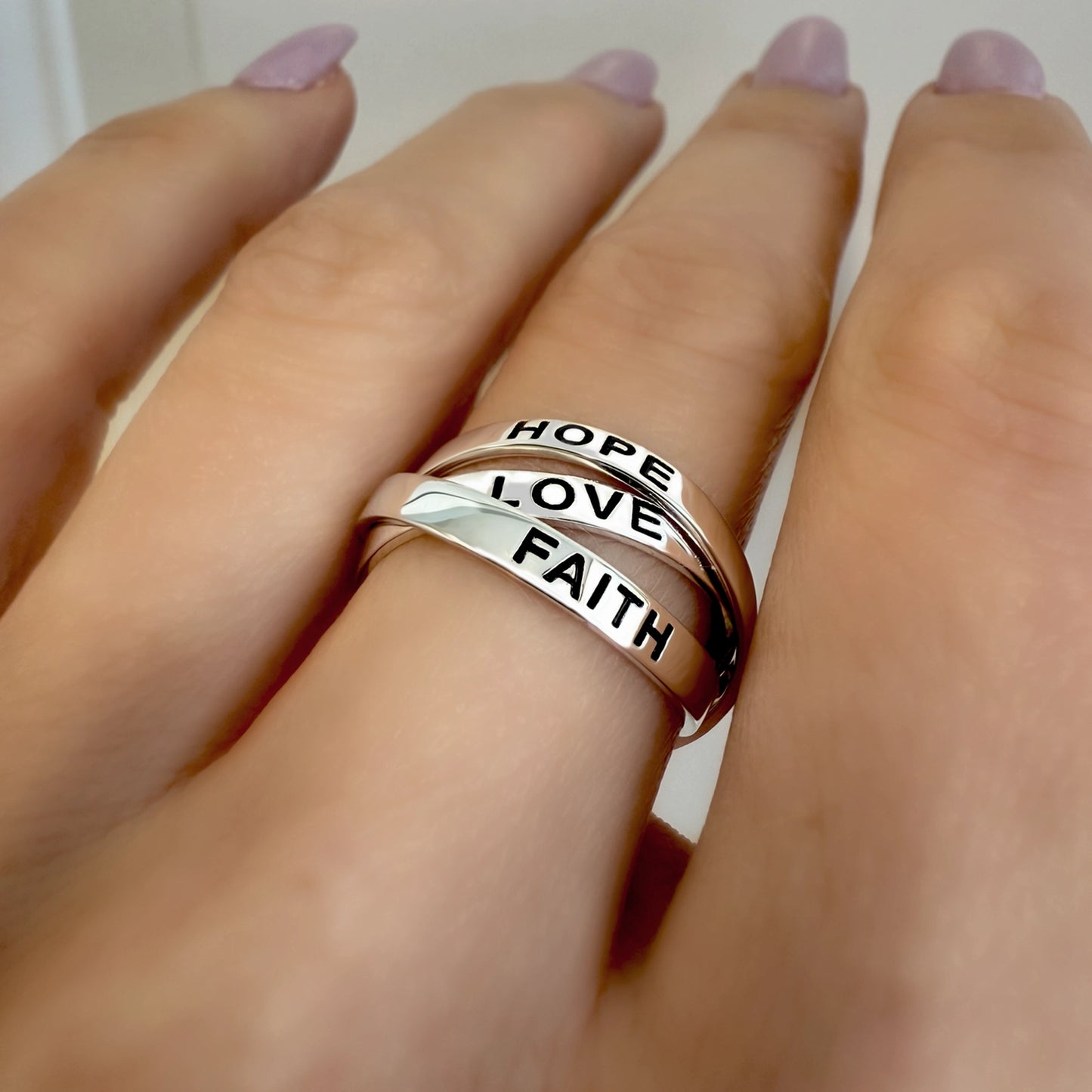 Close-up of a hand wearing a sterling silver triple band ring engraved with the words "FAITH," "LOVE," and "HOPE" on each band, stacked together.