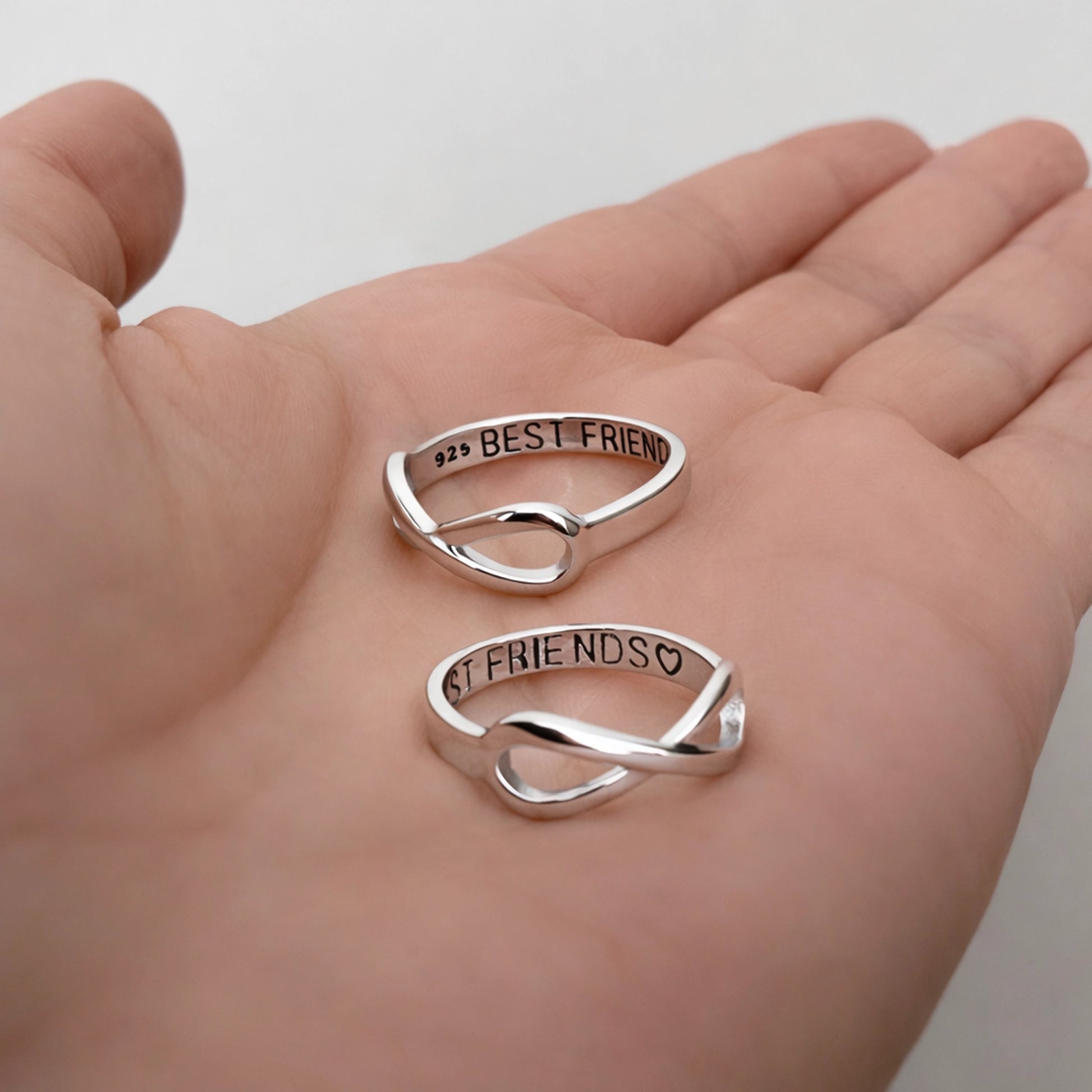 Sterling silver infinity best friend rings resting in a palm, engraved with “BEST FRIEND” and “FRIENDS” with heart, polished finish, minimalist friendship jewelry close-up.