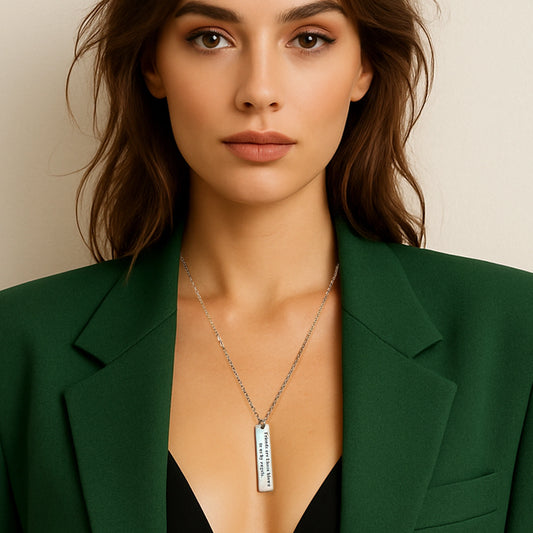 Woman wearing a green blazer and black top showcases a sterling silver tag necklace engraved with the message “Friends are kisses blown to us by angels.” The rectangular pendant hangs from a delicate silver chain, adding a sentimental and elegant touch to her minimalist outfit.