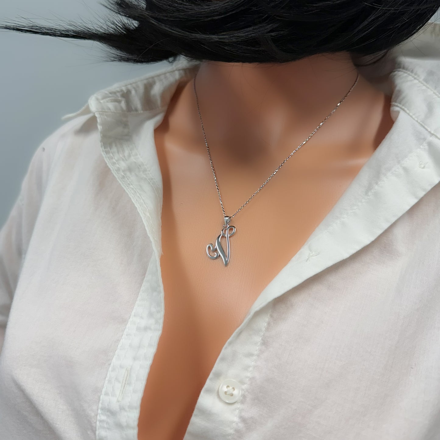 Close-up of a sterling silver initial pendant necklace on a dainty chain, displayed on a model with a white shirt slightly open at the neckline.