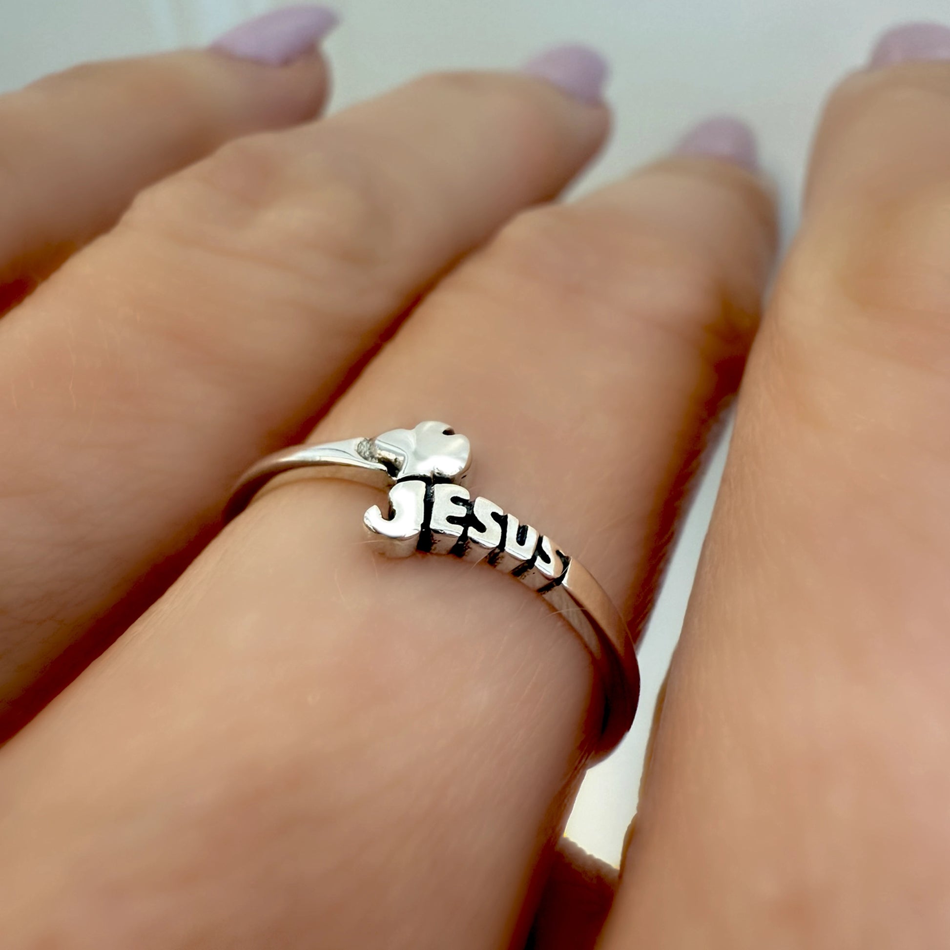 Close-up of a sterling silver chevron ring featuring the word “JESUS” in bold 3D letters and a small dove detail on top, worn on a woman’s finger with light purple polished nails.