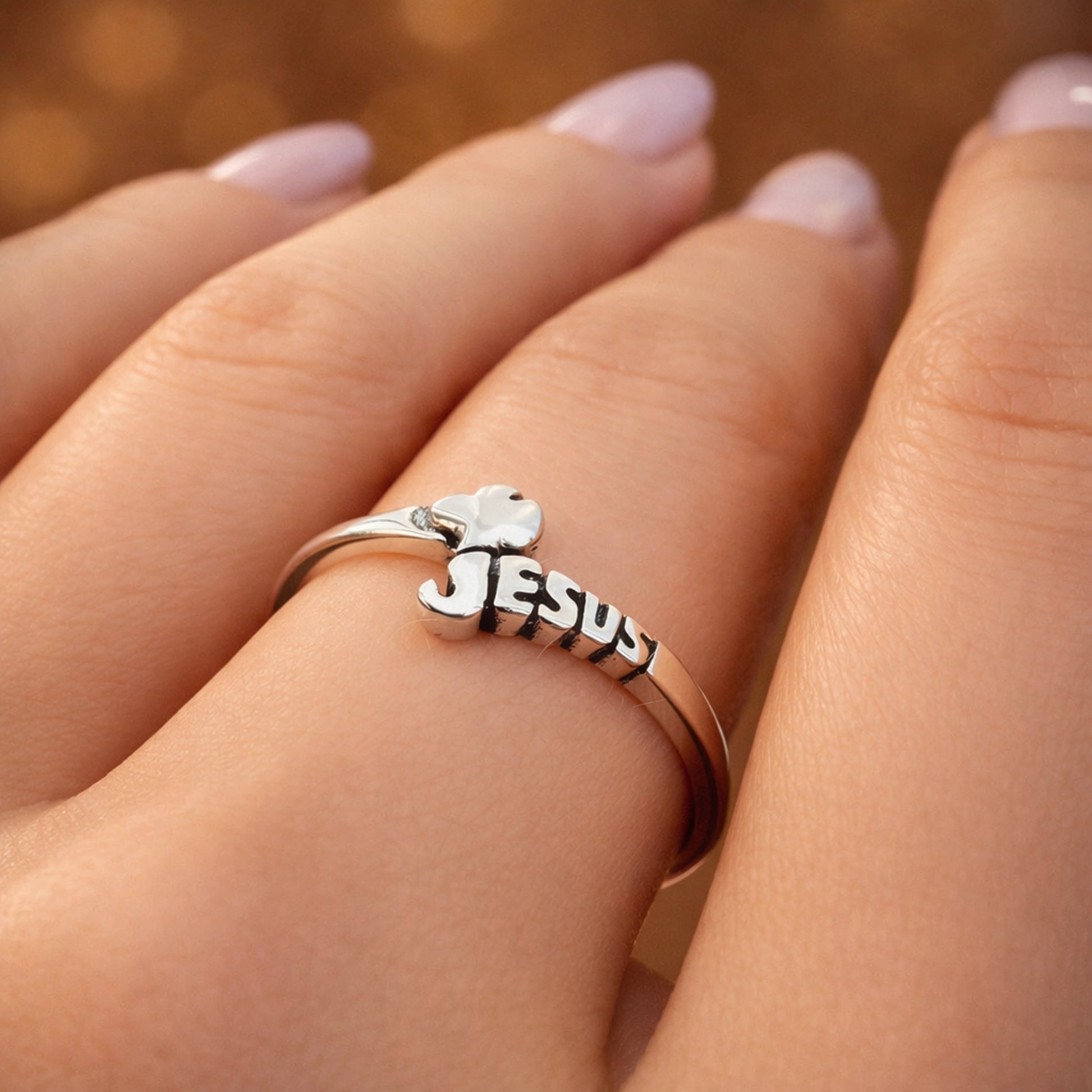 Close-up lifestyle photo of a delicate sterling silver Jesus name ring with a dove worn on a woman's hand. The polished silver band features the word Jesus and a small dove detail, symbolizing Christian faith and devotion. The ring is shown on a model with a soft French manicure, captured with warm studio lighting and a blurred background that highlights the shine and fine details of the religious jewelry.