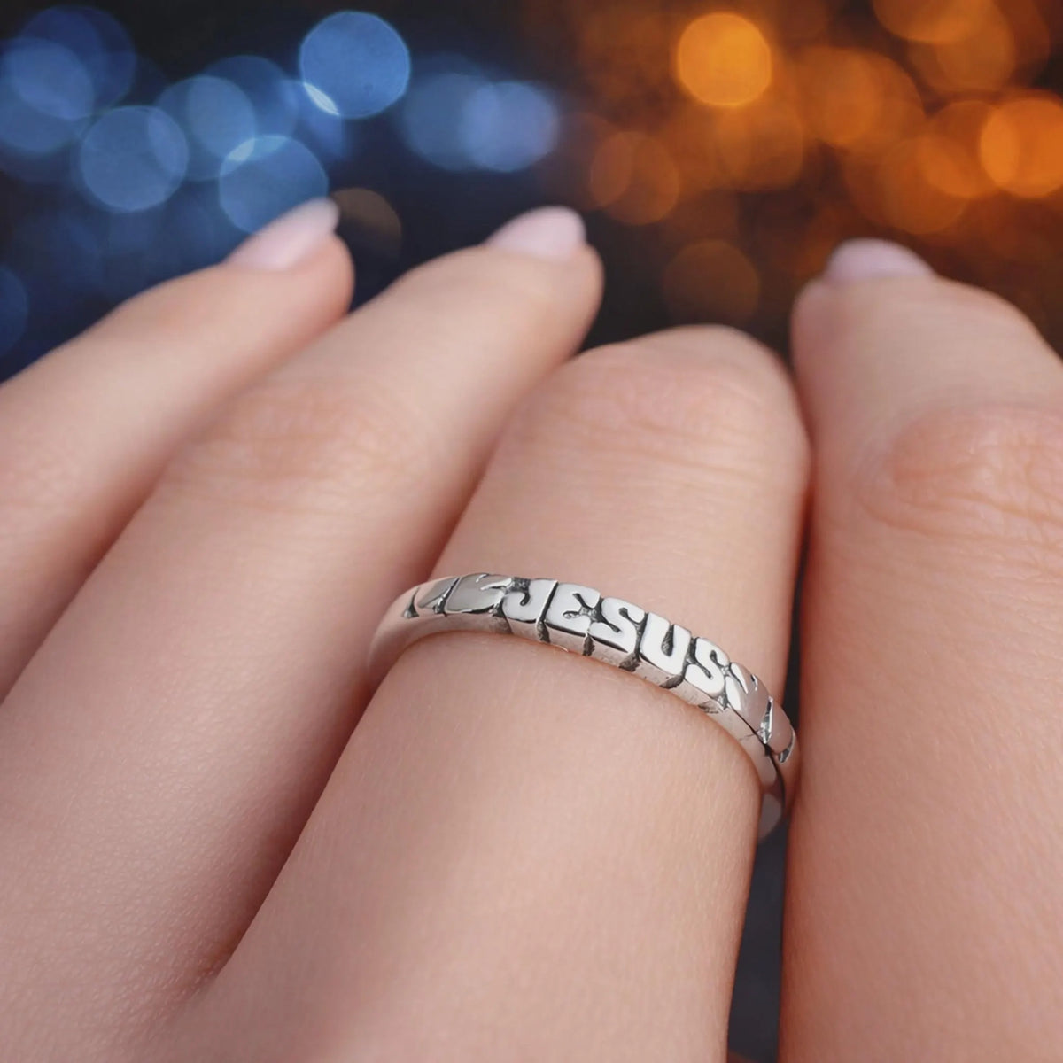 Sterling silver Jesus band ring with cut-out lettering spelling “JESUS,” shown on a hand with light pink manicured nails against a natural green background.