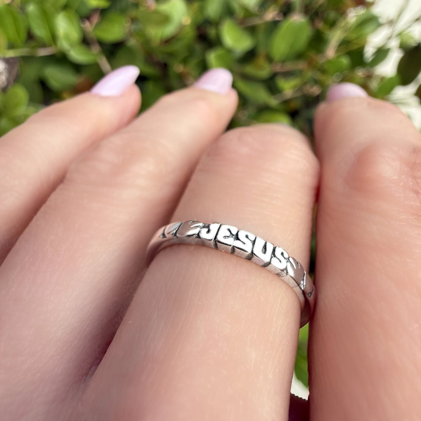 Sterling silver Jesus band ring with cut-out lettering spelling “JESUS,” shown on a hand with light pink manicured nails against a natural green background.
