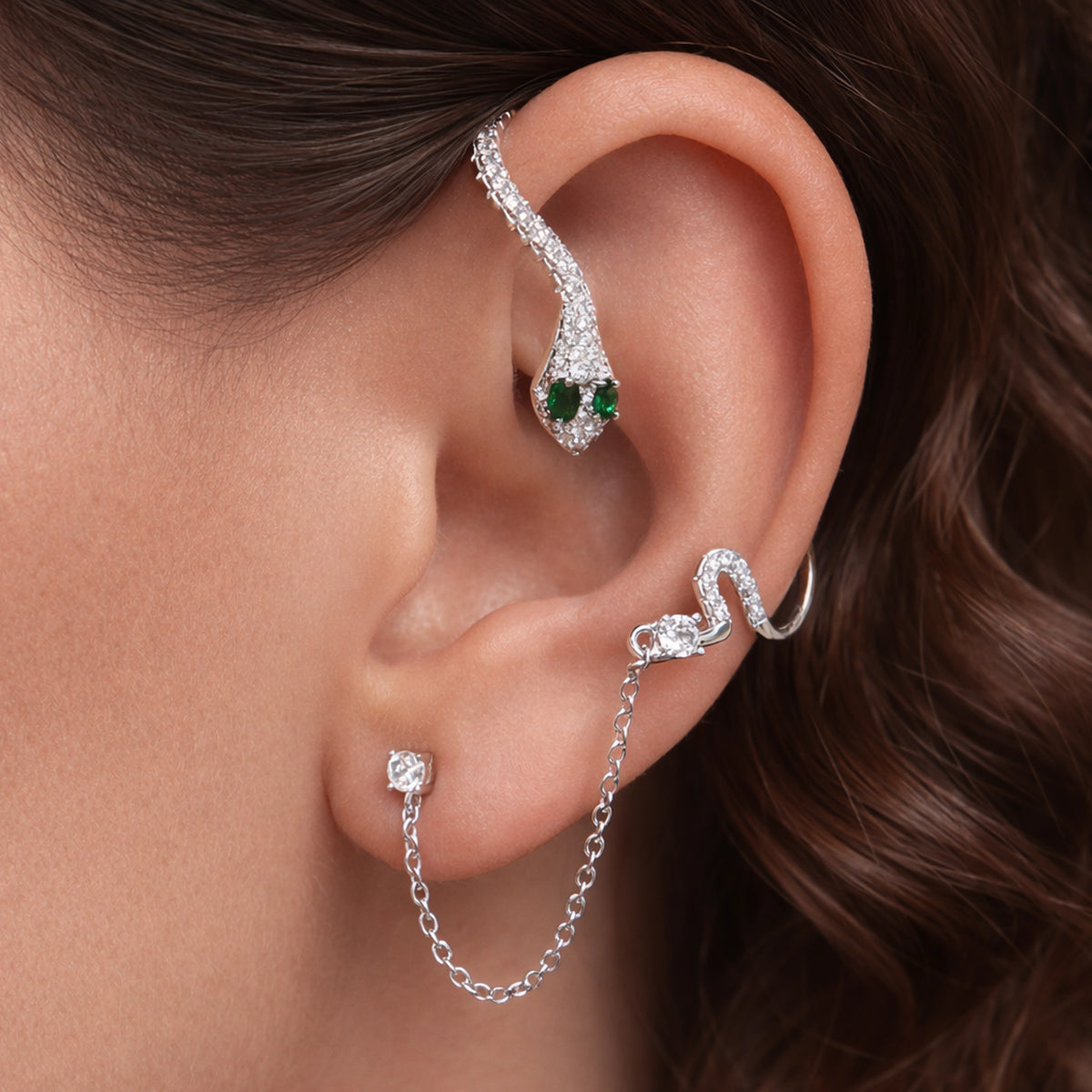 Silver snake ear cuff with emerald green eyes and sparkling pavé crystals on a model’s ear, styled with delicate silver chain link stud earring -  edgy statement ear stack jewelry close-up.