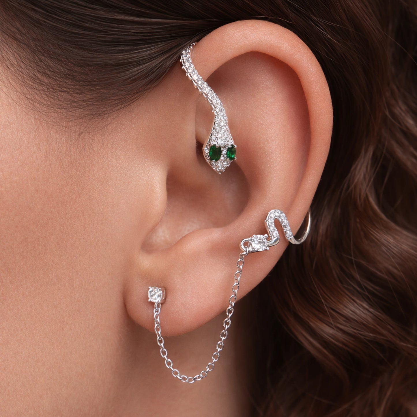 Silver snake ear cuff with emerald green eyes and sparkling pavé crystals on a model’s ear, styled with delicate silver chain link stud earring -  edgy statement ear stack jewelry close-up.