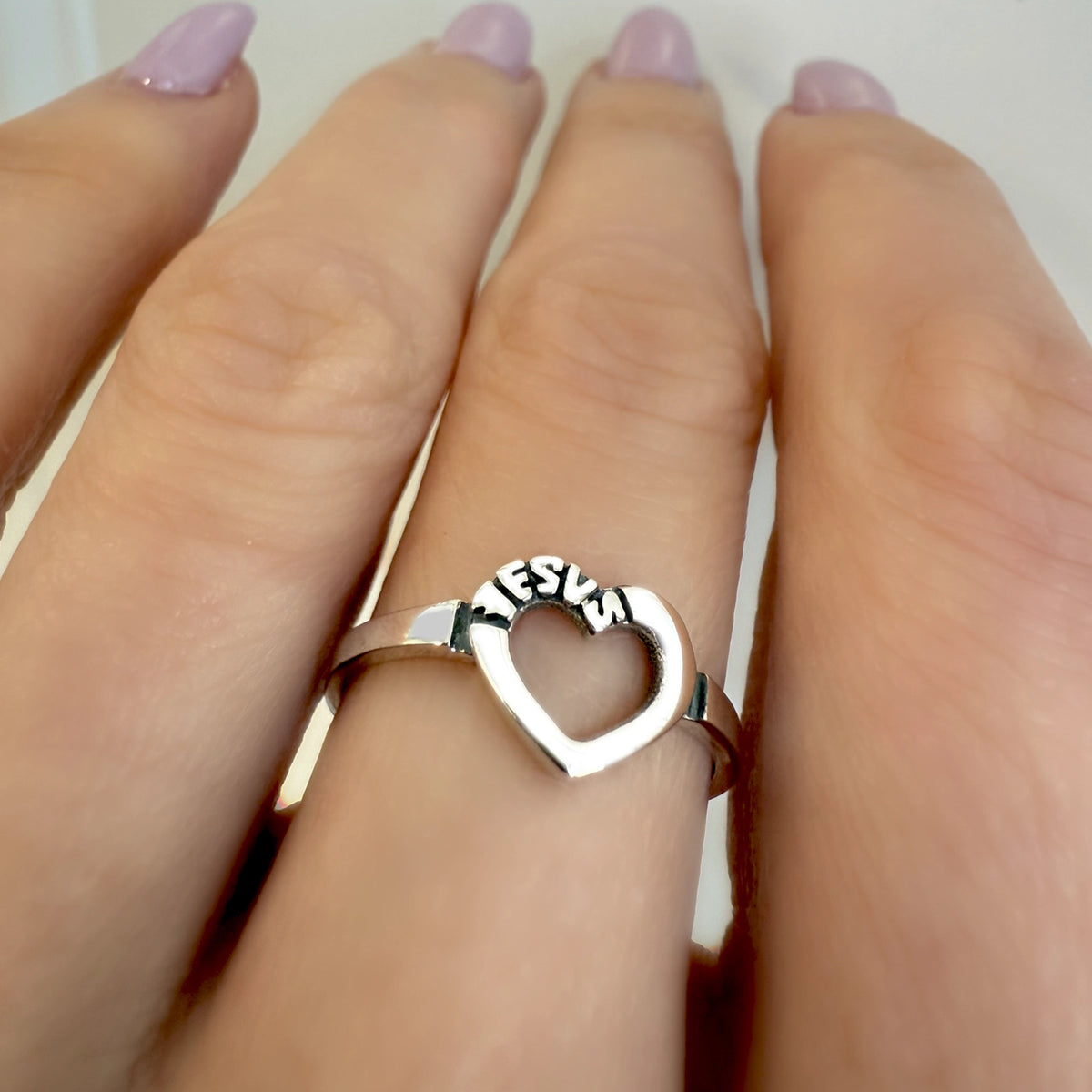 Sterling silver open heart ring engraved with the word "JESUS," worn on a woman's finger with light lavender manicure.