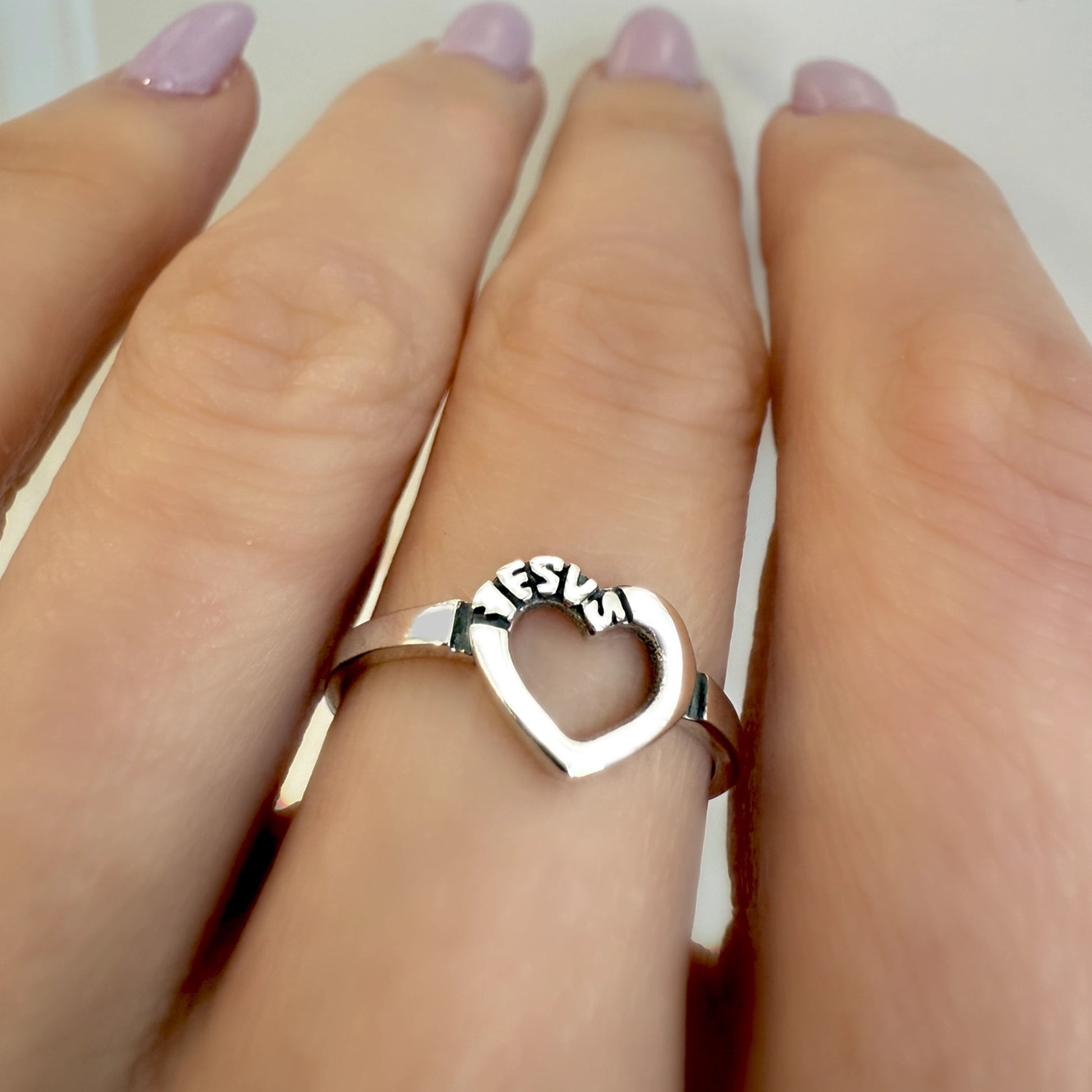 Sterling silver open heart ring engraved with the word "JESUS," worn on a woman's finger with light lavender manicure.