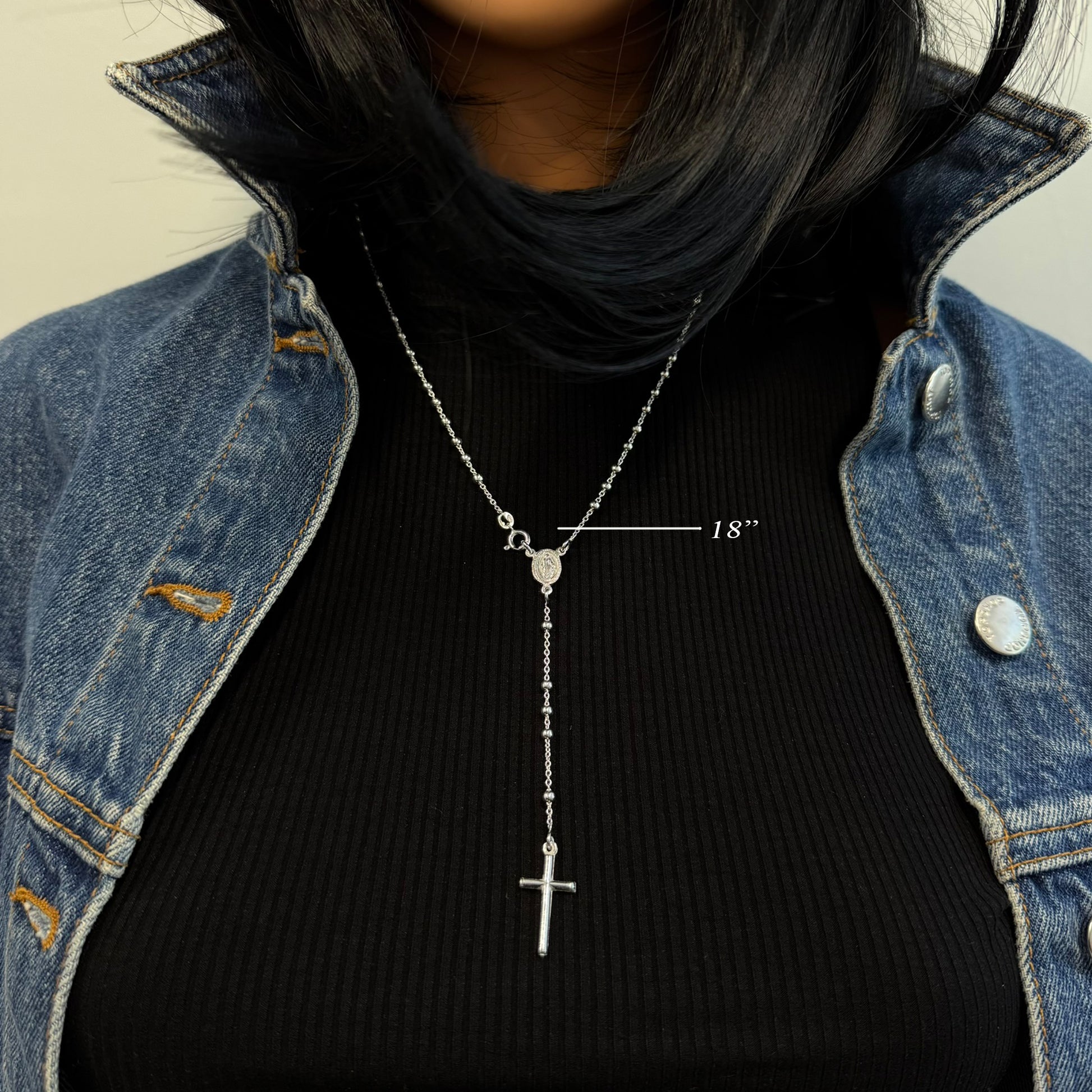 Model wearing an 18-inch sterling silver rosary bead necklace with a Virgin Mary connector and hanging cross pendant. Styled over a black ribbed top and denim jacket for a casual layered look.