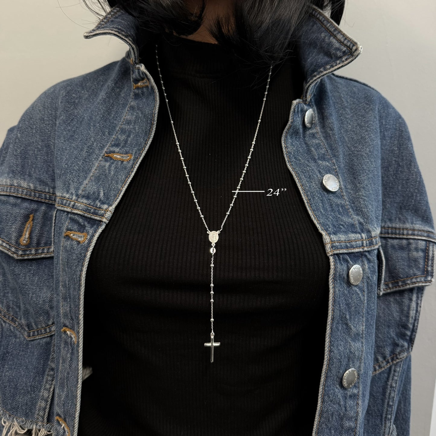Model wearing a 24-inch sterling silver rosary necklace with small round beads, Virgin Mary centerpiece, and vertical cross pendant. Styled with a black ribbed shirt and open denim jacket.