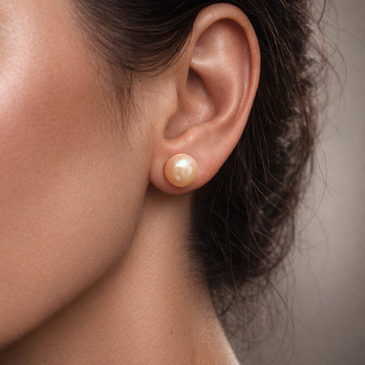 Peach pearl stud earring on model’s ear, round glossy freshwater pearl with soft warm luster, minimalist elegant jewelry, classic bridal accessory, close-up studio lighting, neutral background.