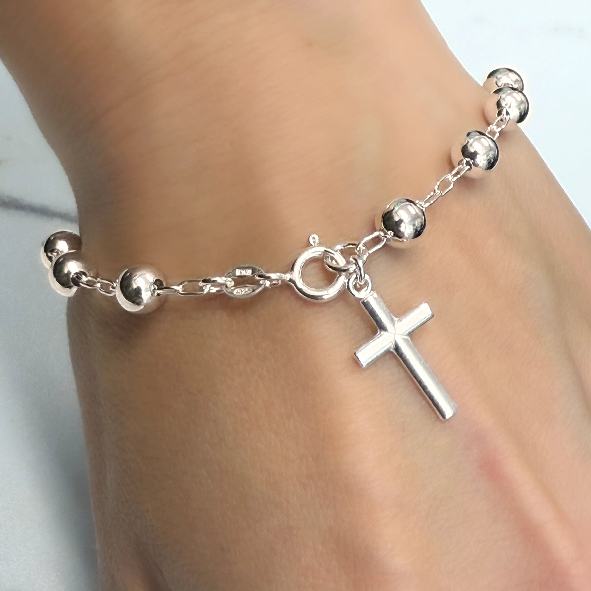 Sterling silver beaded bracelet featuring a delicate chain with shiny round ball accents and a small vertical cross charm. The bracelet is secured with a spring ring clasp and worn on a woman's wrist against a marble surface.