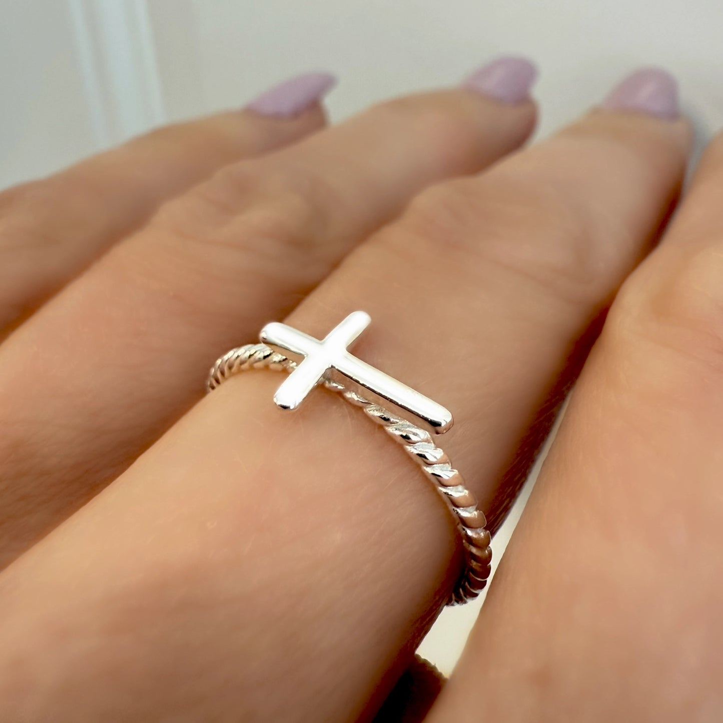 Sterling silver ring featuring a horizontal cross on a twisted rope-style band, elegantly worn on a woman's finger with pastel pink manicure.