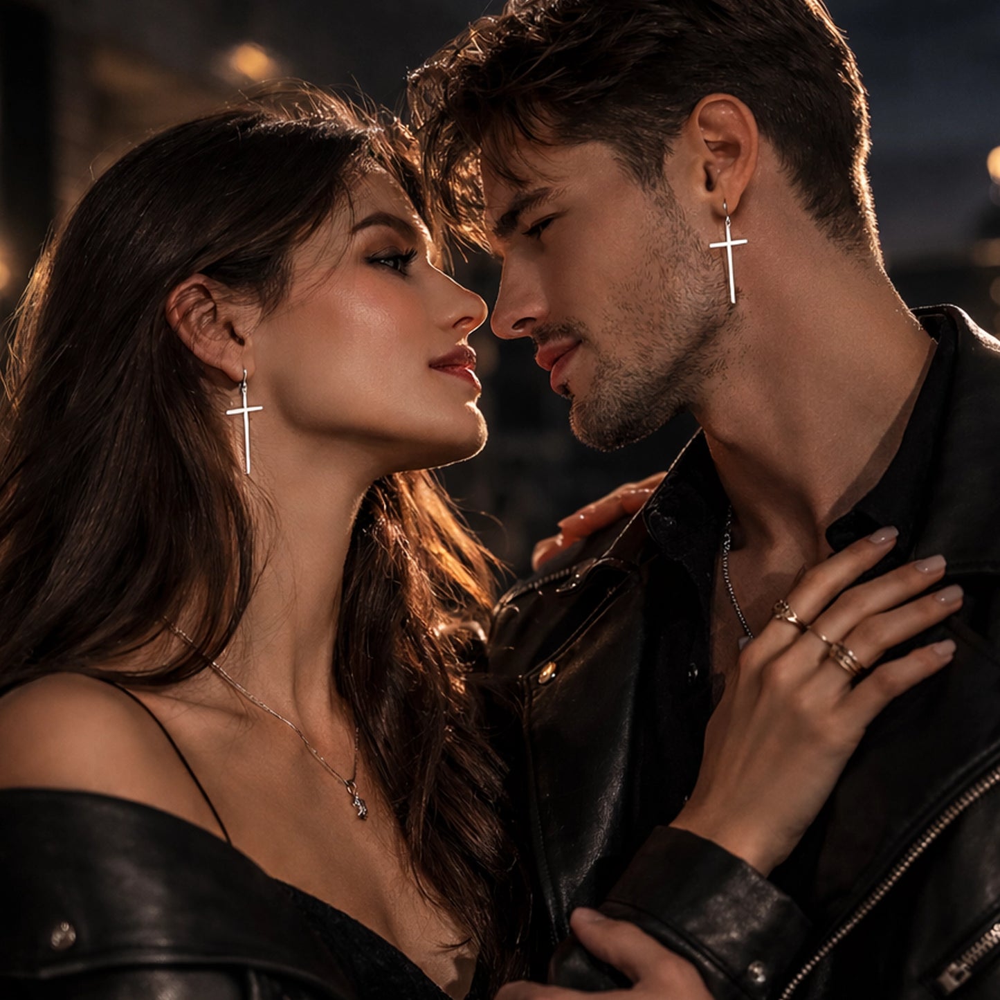 Silver cross dangle earrings worn by stylish couple in romantic lifestyle portrait, close-up man and woman facing each other, wearing matching minimalist sterling silver cross drop earrings, leather jackets, cinematic dark background with warm bokeh lights, modern fashion jewelry editorial style.