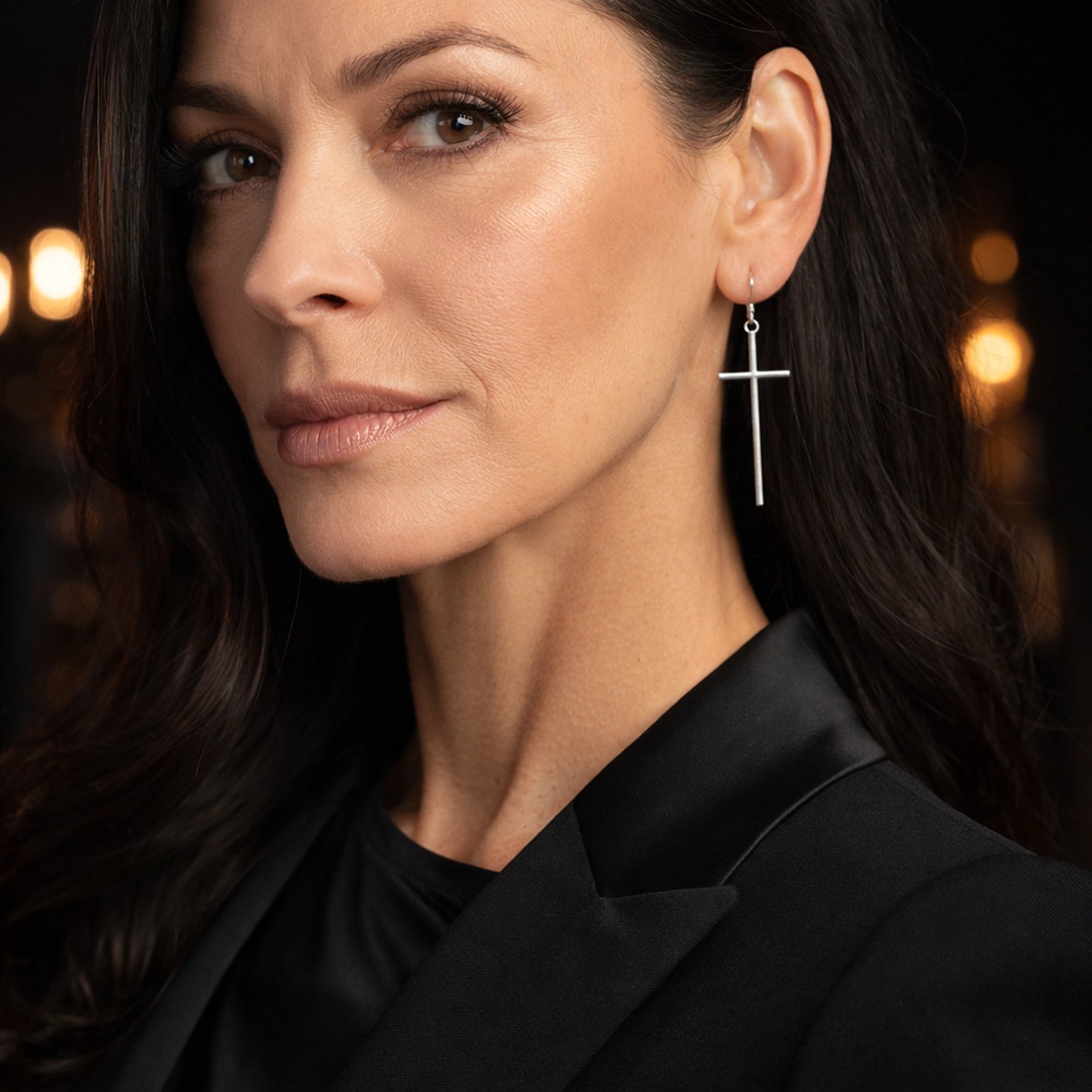 Close-up portrait of a stylish, well-groomed woman around 55–60 wearing a silver cross dangle earring, shown in sharp detail against a softly blurred warm studio background.