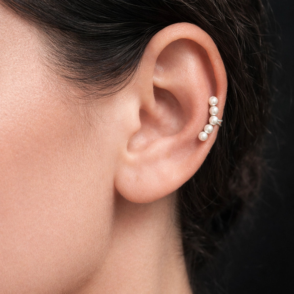Close-up of a woman’s ear wearing a sterling silver single dainty pearl ear cuff with small round pearls aligned along the cartilage. Minimalist elegant ear jewelry styled on smooth skin with soft professional lighting, highlighting the delicate pearl ear climber design.