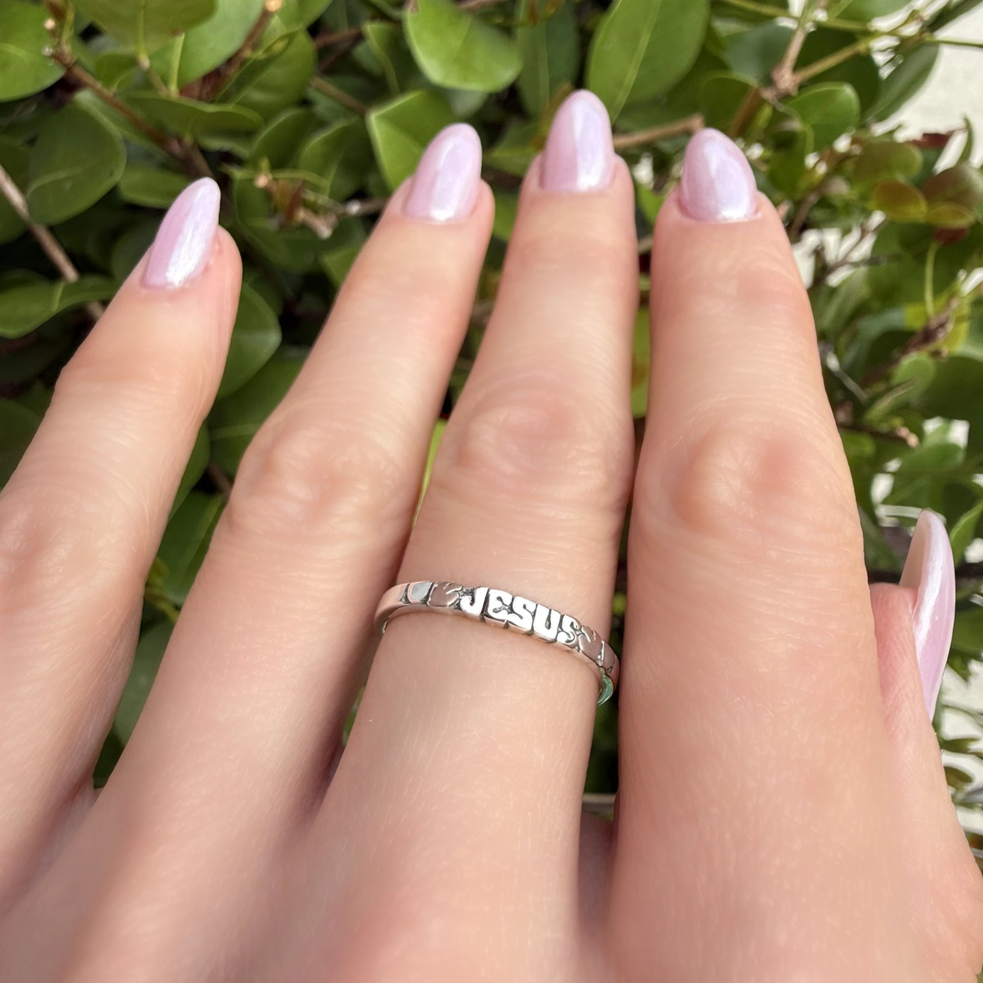 Sterling silver tiny Jesus band ring on hand, engraved with the word ‘JESUS’ in bold cutout letters, styled with soft pink manicure and green leaf background.