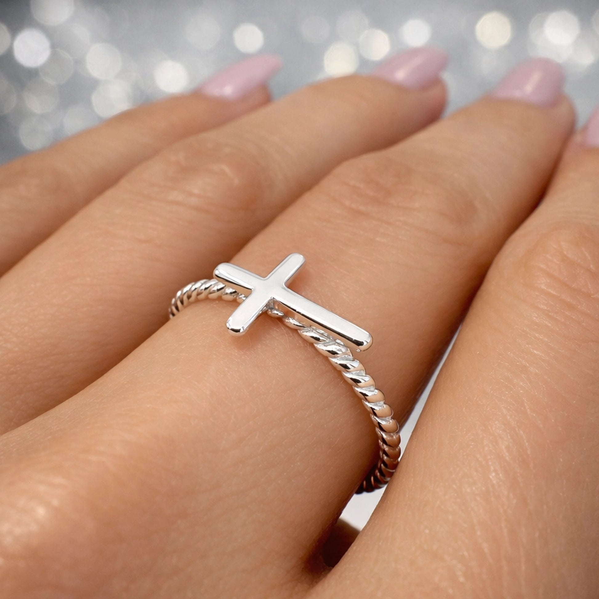Sterling silver ring featuring a horizontal cross on a twisted rope-style band, elegantly worn on a woman's finger with pastel pink manicure.