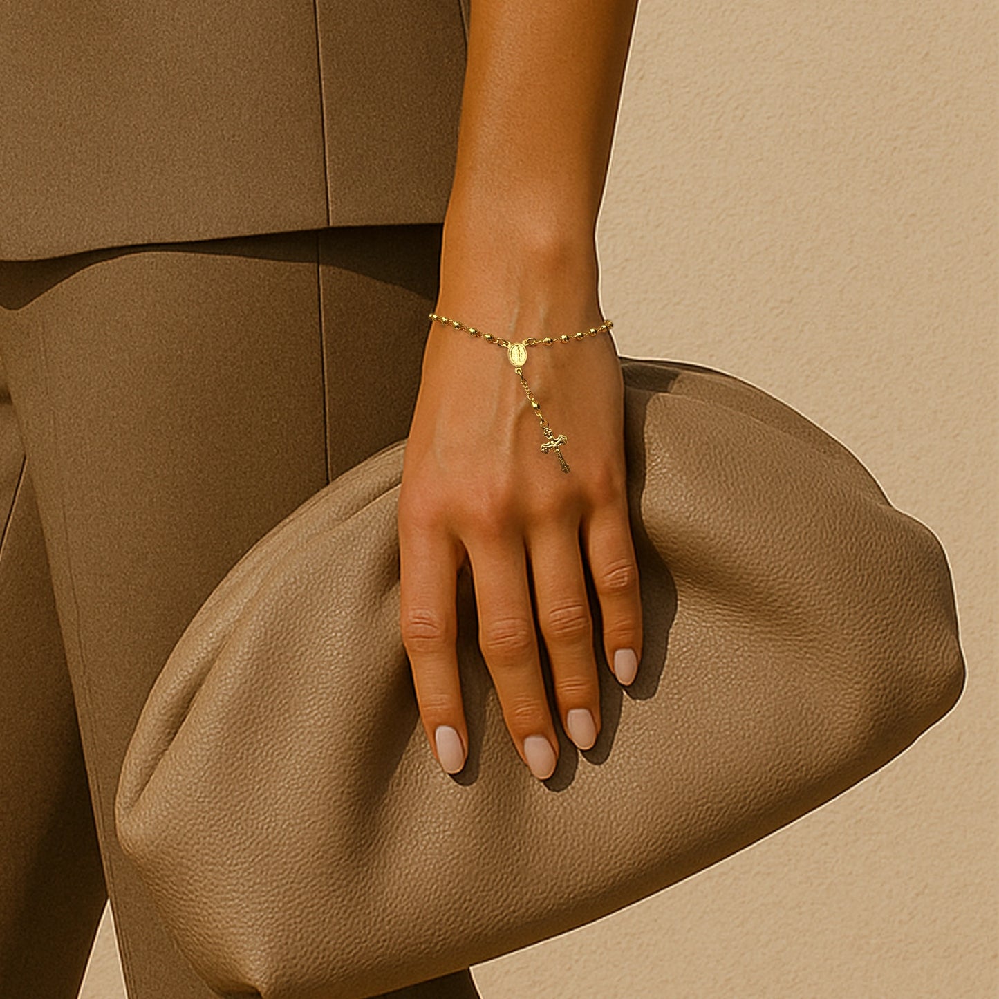 Faith-inspired gold rosary bead bracelet with Virgin Mary charm and dangling cross, worn elegantly with taupe outfit.