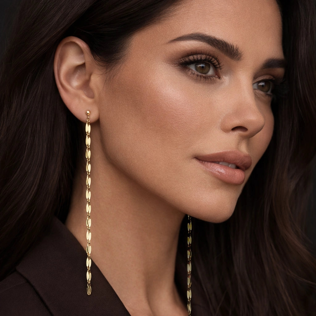 Beautiful model wearing extra long 18K gold filled chain drop earrings with oval textured links, both ears visible. Elegant studio portrait, glossy makeup, flawless skin, voluminous dark hair, chocolate brown oversized blazer. Luxury minimalist jewelry, high contrast, professional lighting, square format.