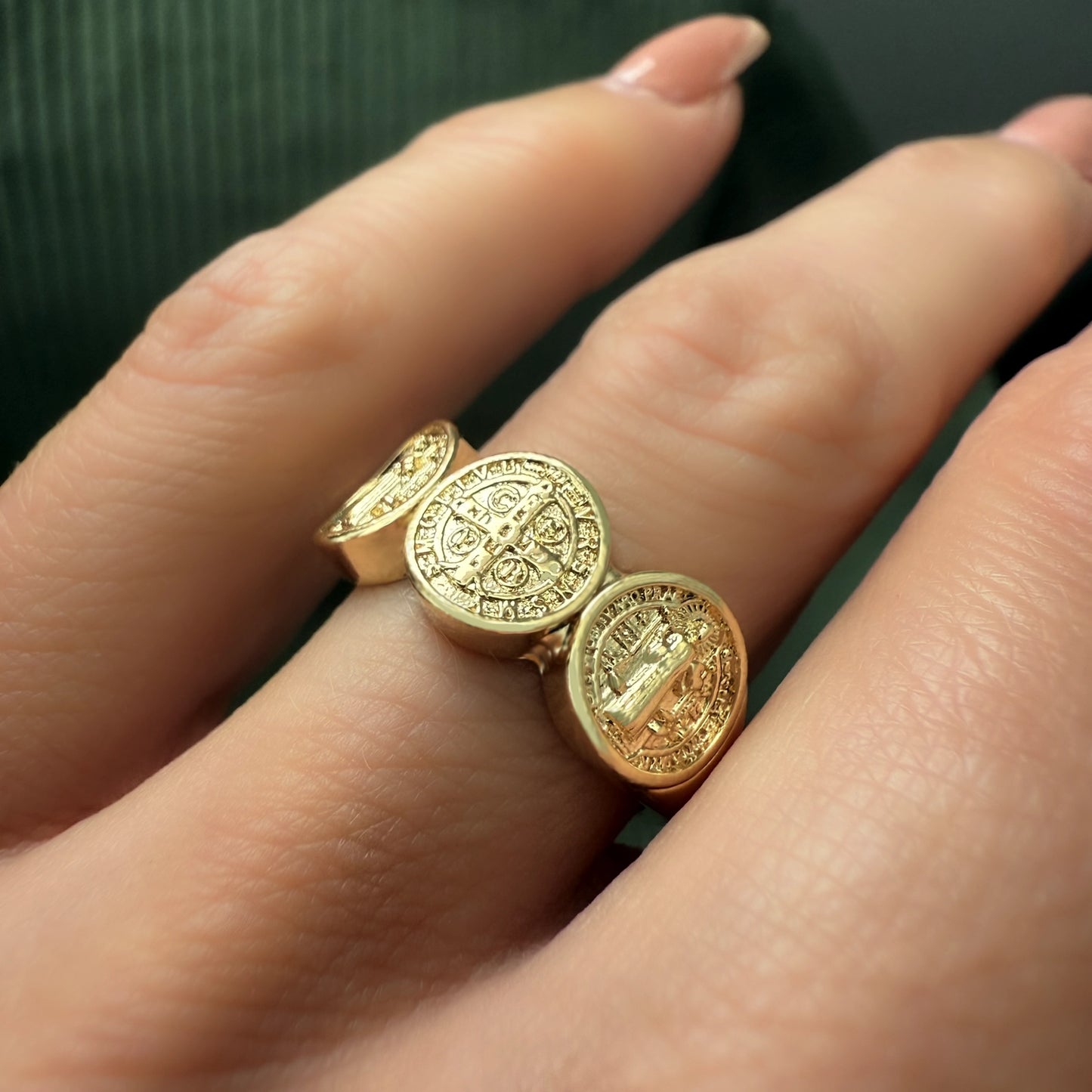Close-up of a woman's hand wearing a gold filled triple medallion ring featuring Saint Benedict and cross designs, showcasing the intricate religious engravings on each oval face against a dark green background.