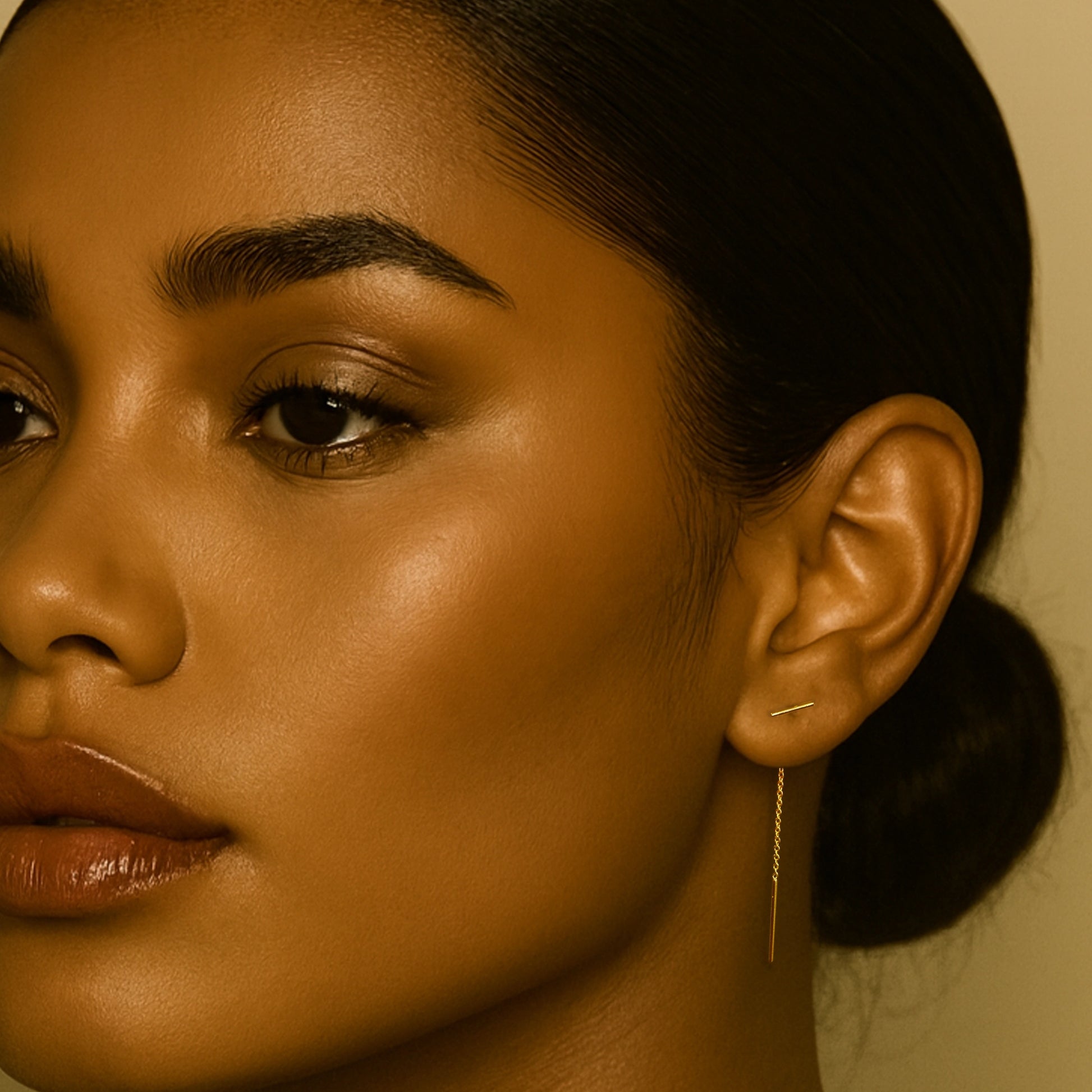 Close-up of a woman with glowing deep skin tone wearing minimal makeup, showcasing a delicate gold threader earring with a tiny bar design.